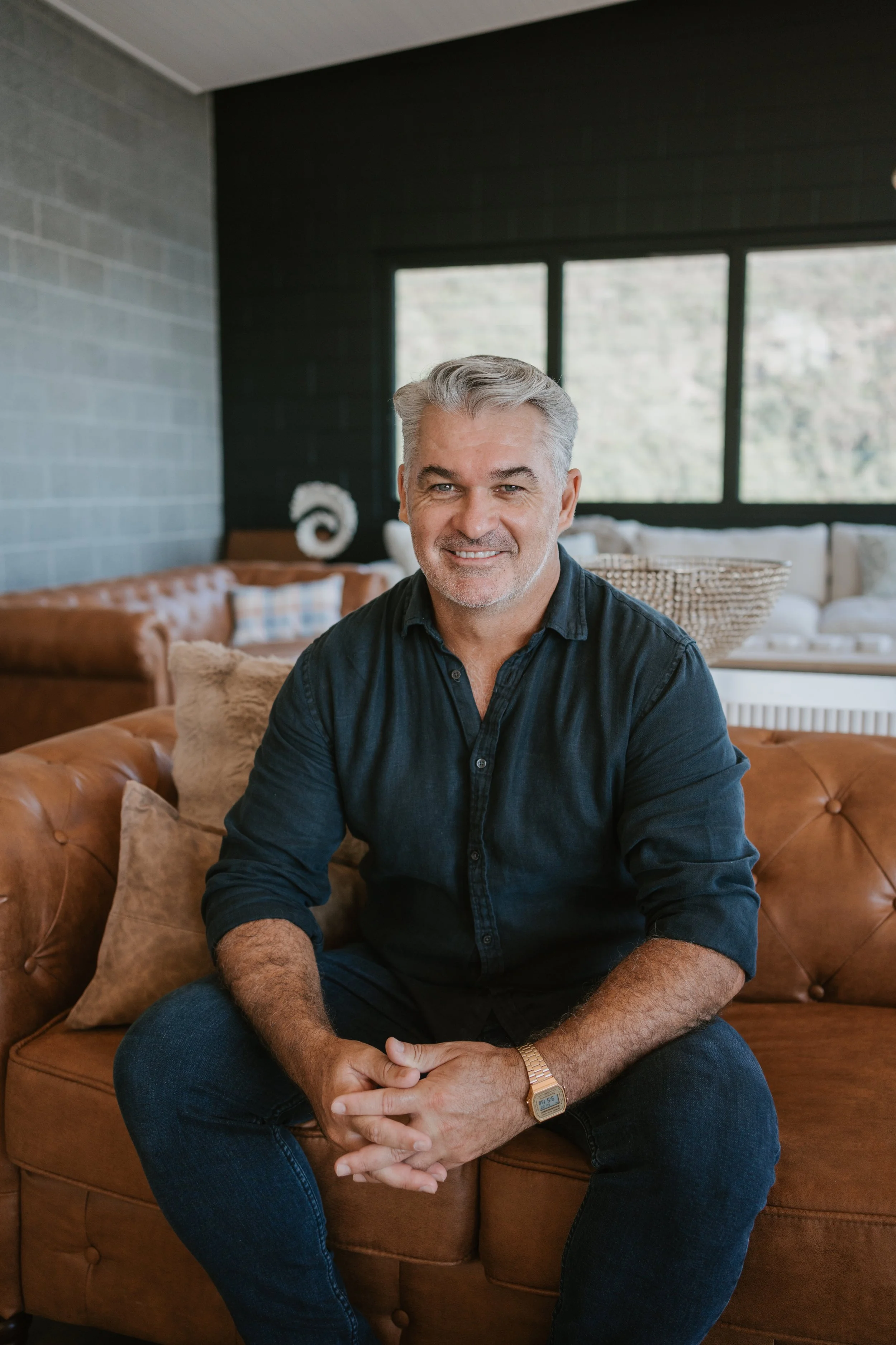 A middle-aged man with gray hair and a beard, wearing a dark button-up shirt, sitting on a brown leather sofa in a modern living room with large windows, a black wall, and decorative pillows.