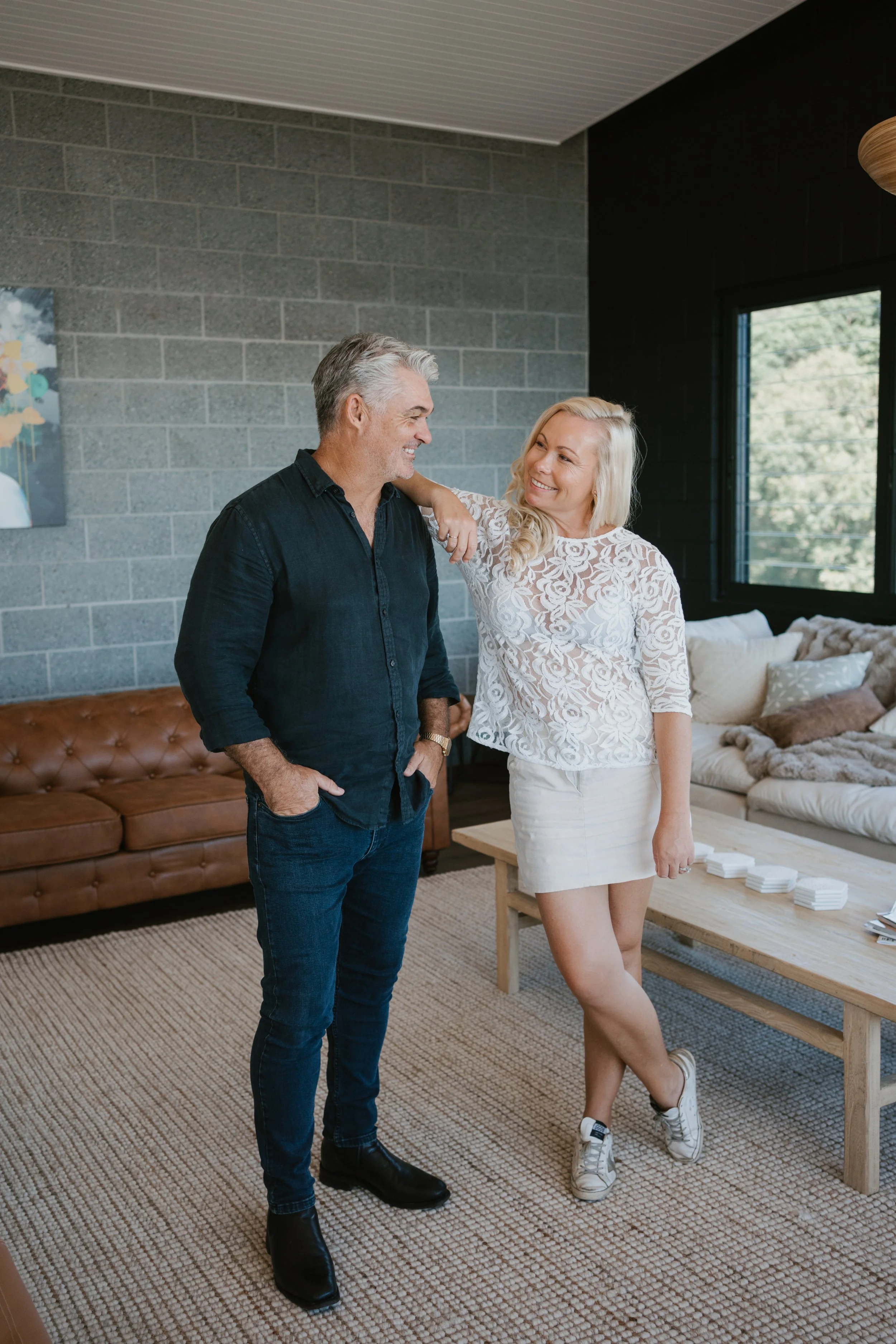 A man and woman standing inside a modern living room, smiling and looking at each other, with the woman resting her arm on the man's shoulder.
