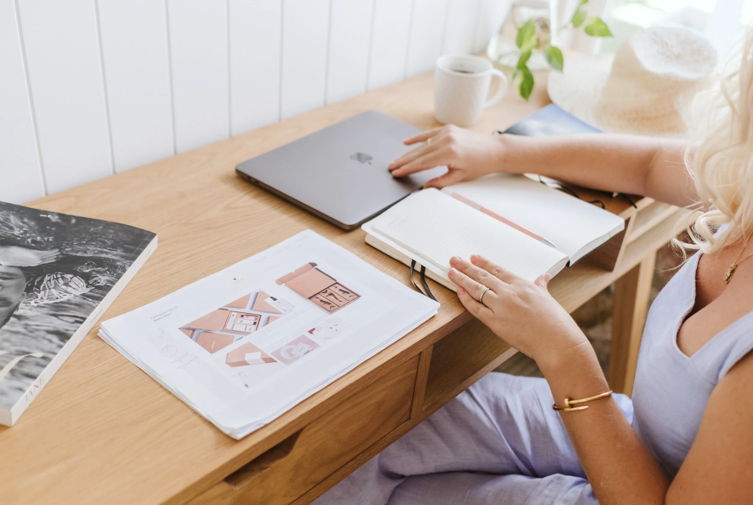 Person sitting at a wooden desk with a closed laptop, an open notebook, a pen, printed design plans, and a cup of coffee or tea.