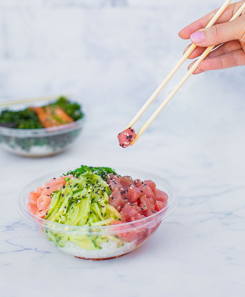 a hand holding a pair of chopstick over a poke bowl containing fish, cucumbers, rice, and seaweed