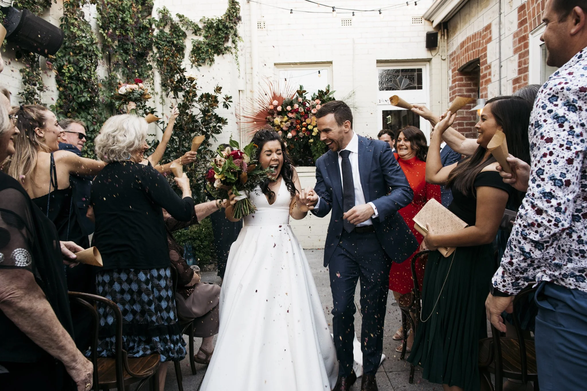 Wedding celebration with a bride in a white gown and a groom in a navy suit holding hands, surrounded by friends and family, some throwing confetti and blowing horns, in an outdoor courtyard with brick and white walls and stringlights.