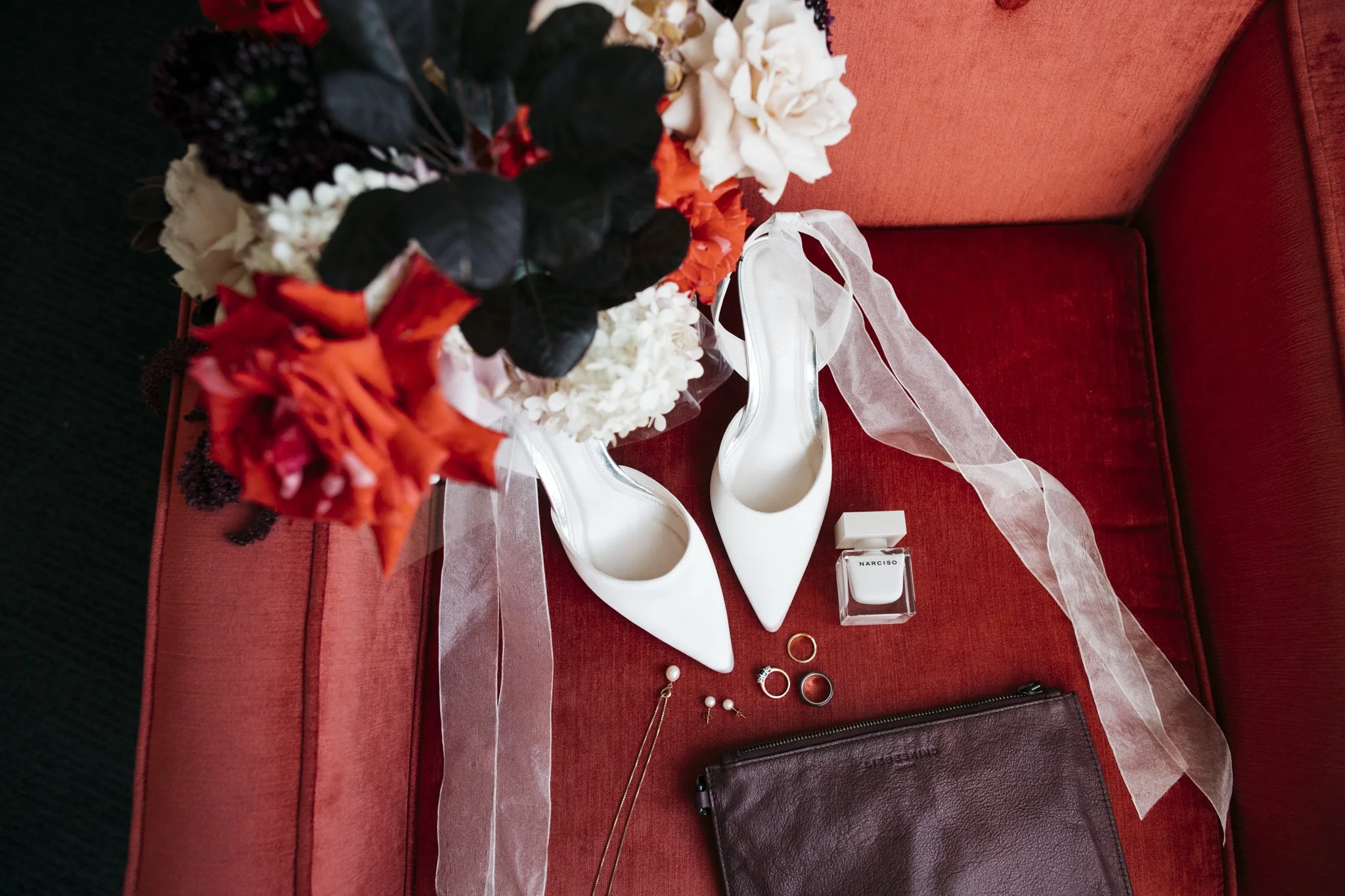 A collection of wedding accessories including white high heels, a vase with red, white, and purple flowers, a bottle of Narciso perfume, gold rings, pearl earrings, a gold necklace, and a brown leather clutch bag arranged on a red cushioned surface.