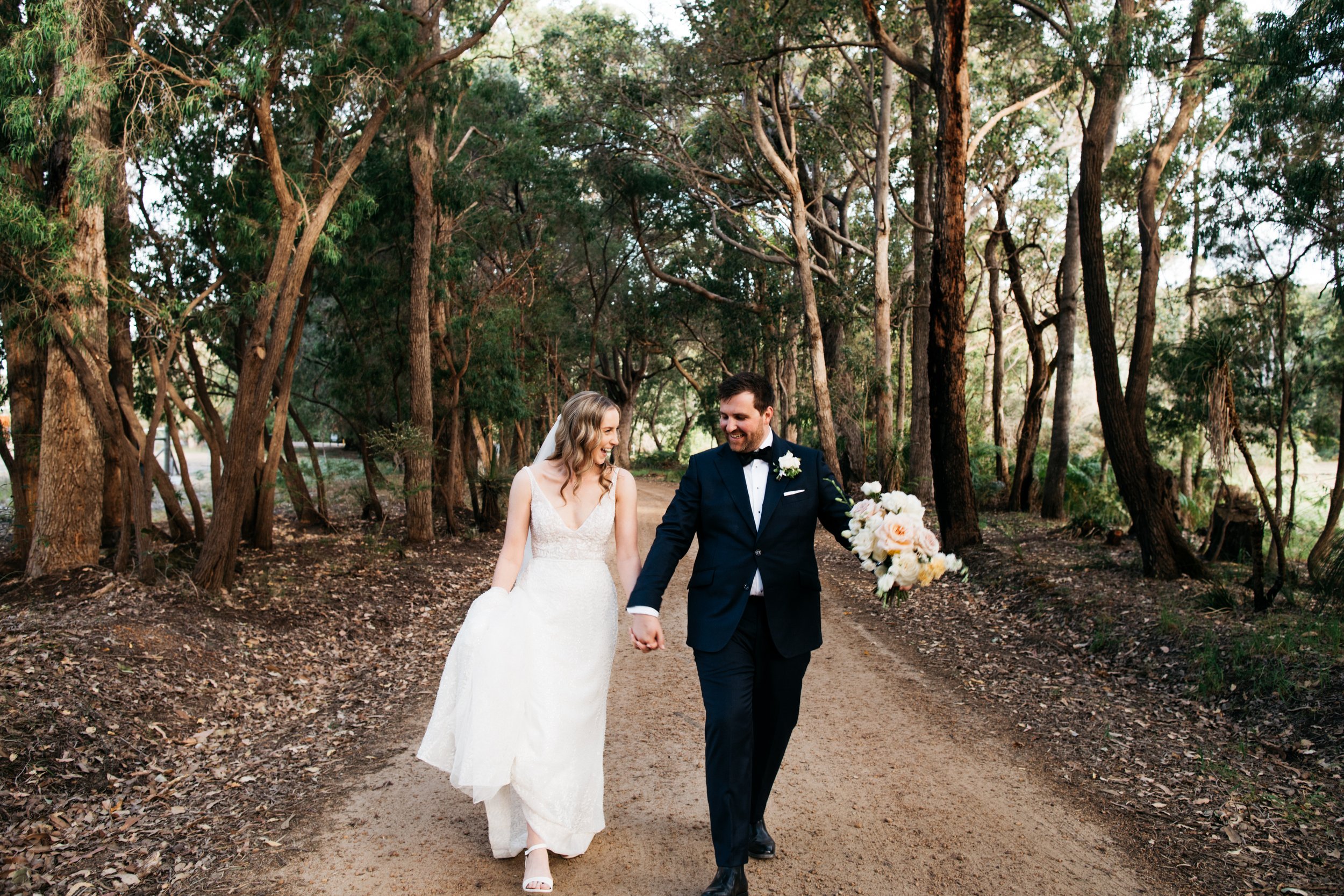 Bride and groom walking hand-in-hand on a forest trail, smiling, with trees and greenery surrounding them, during their wedding.