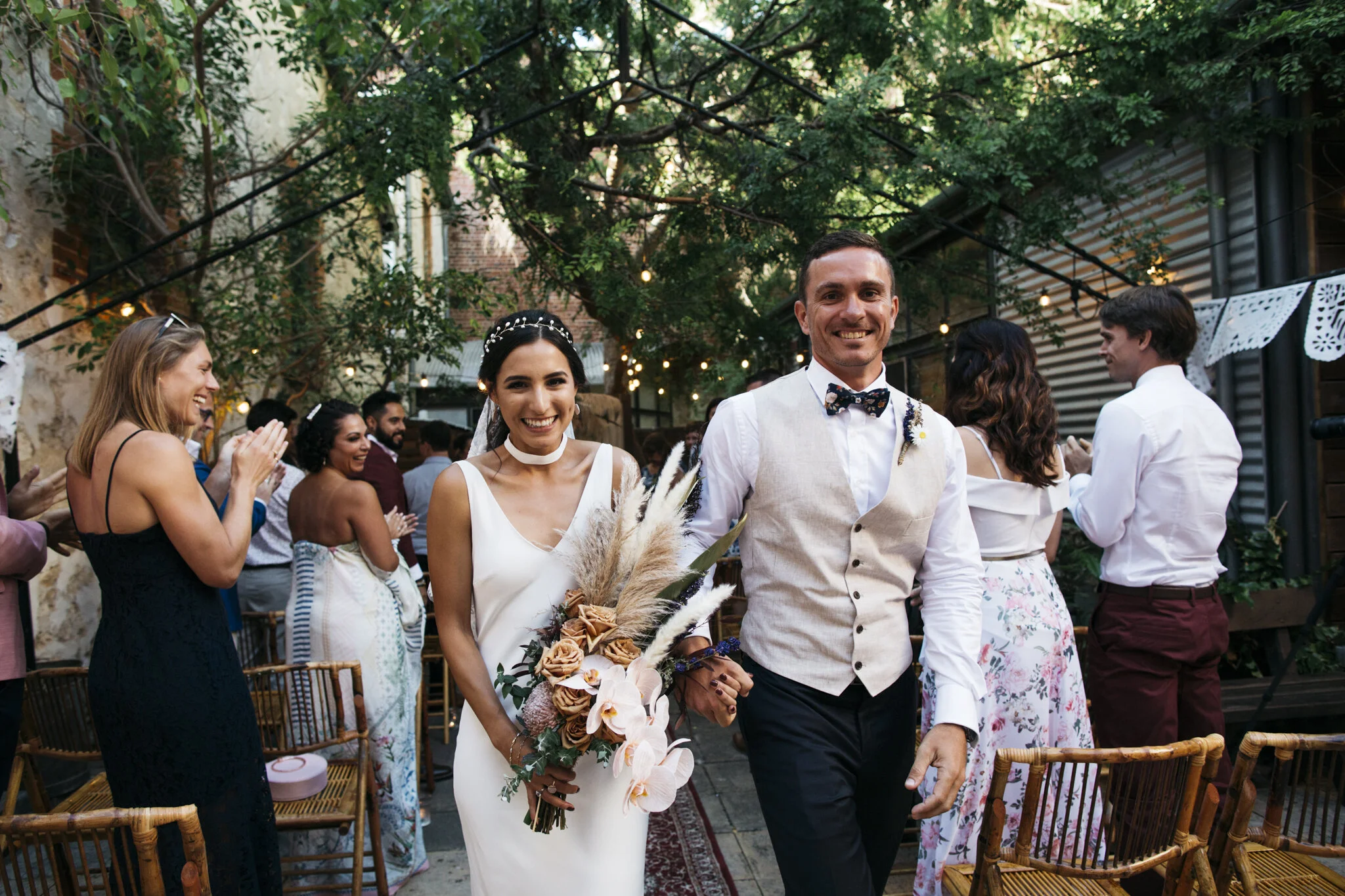 Bride and groom walking arm-in-arm at their outdoor wedding reception, smiling, surrounded by seated and standing guests clapping and celebrating.