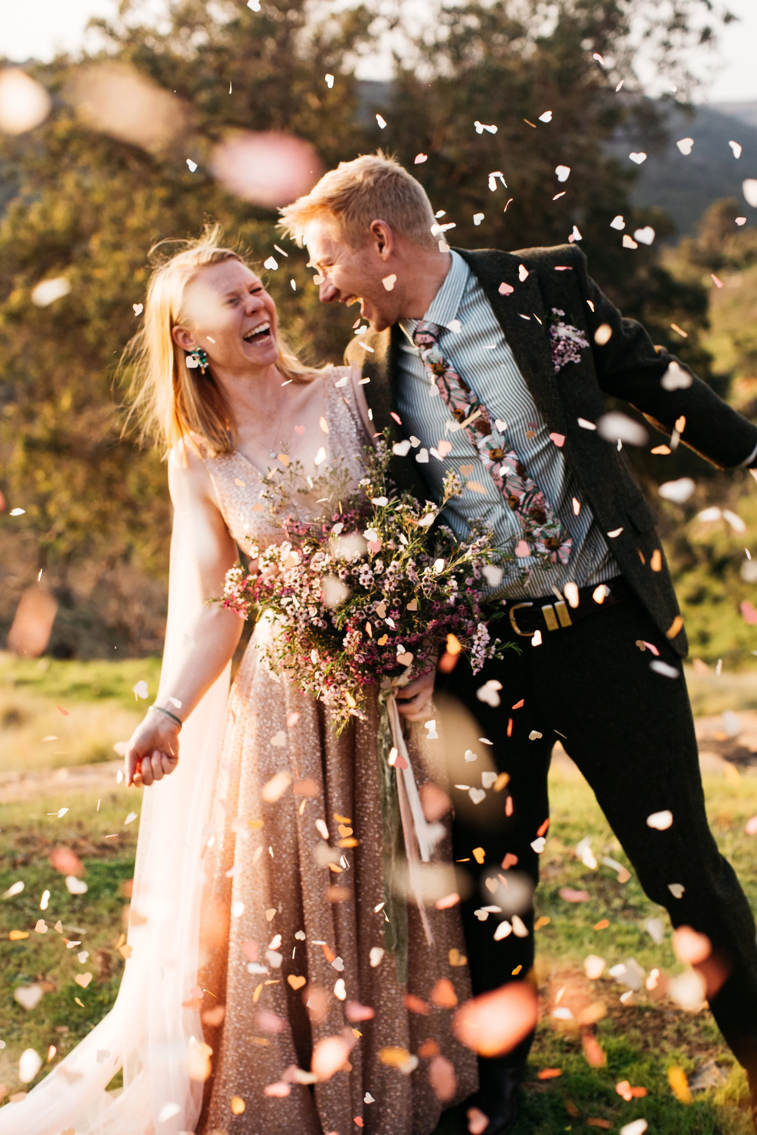 A newlywed couple celebrating outdoors with confetti falling around them, smiling and laughing, with the bride holding a bouquet of flowers.