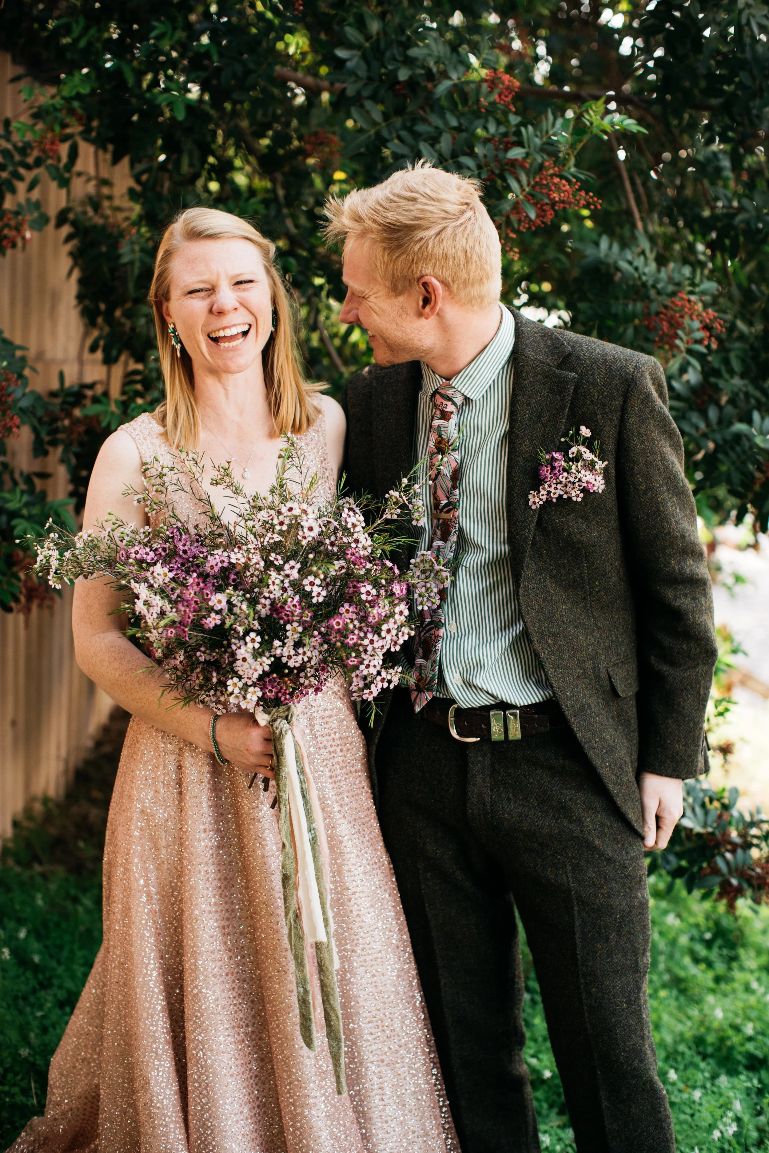 A woman in a pink sequin dress holding a large bouquet of flowers, smiling from laughter. A man in a dark suit and striped shirt is standing next to her, leaning in with an affectionate smile. They are outdoors, surrounded by greenery and flowering bushes.
