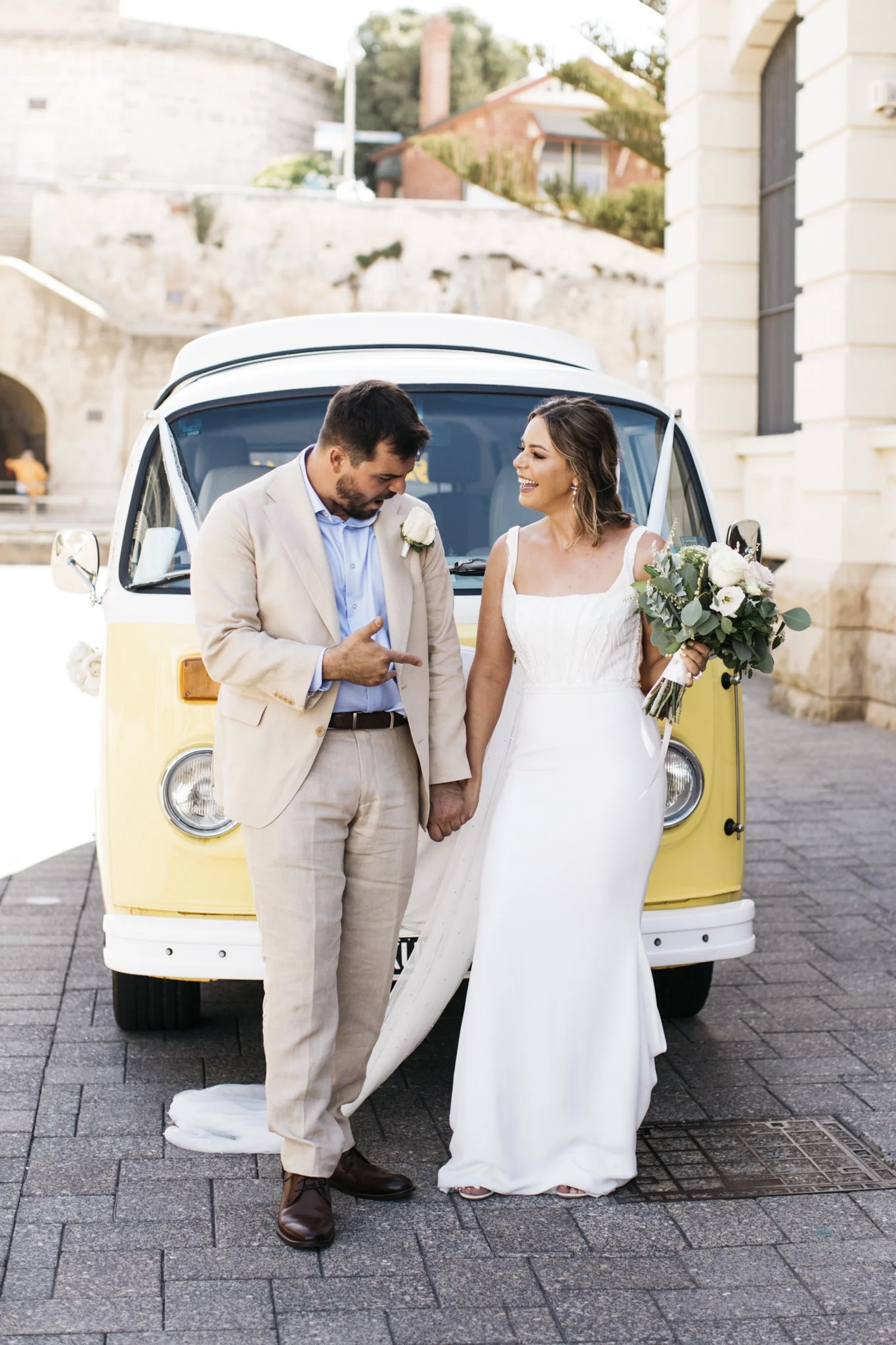 A newlywed couple holding hands and smiling, standing in front of a yellow and white vintage van, with the bride holding a bouquet of flowers. The groom is pointing at the bride's chest area, and they are outdoors in an urban setting.