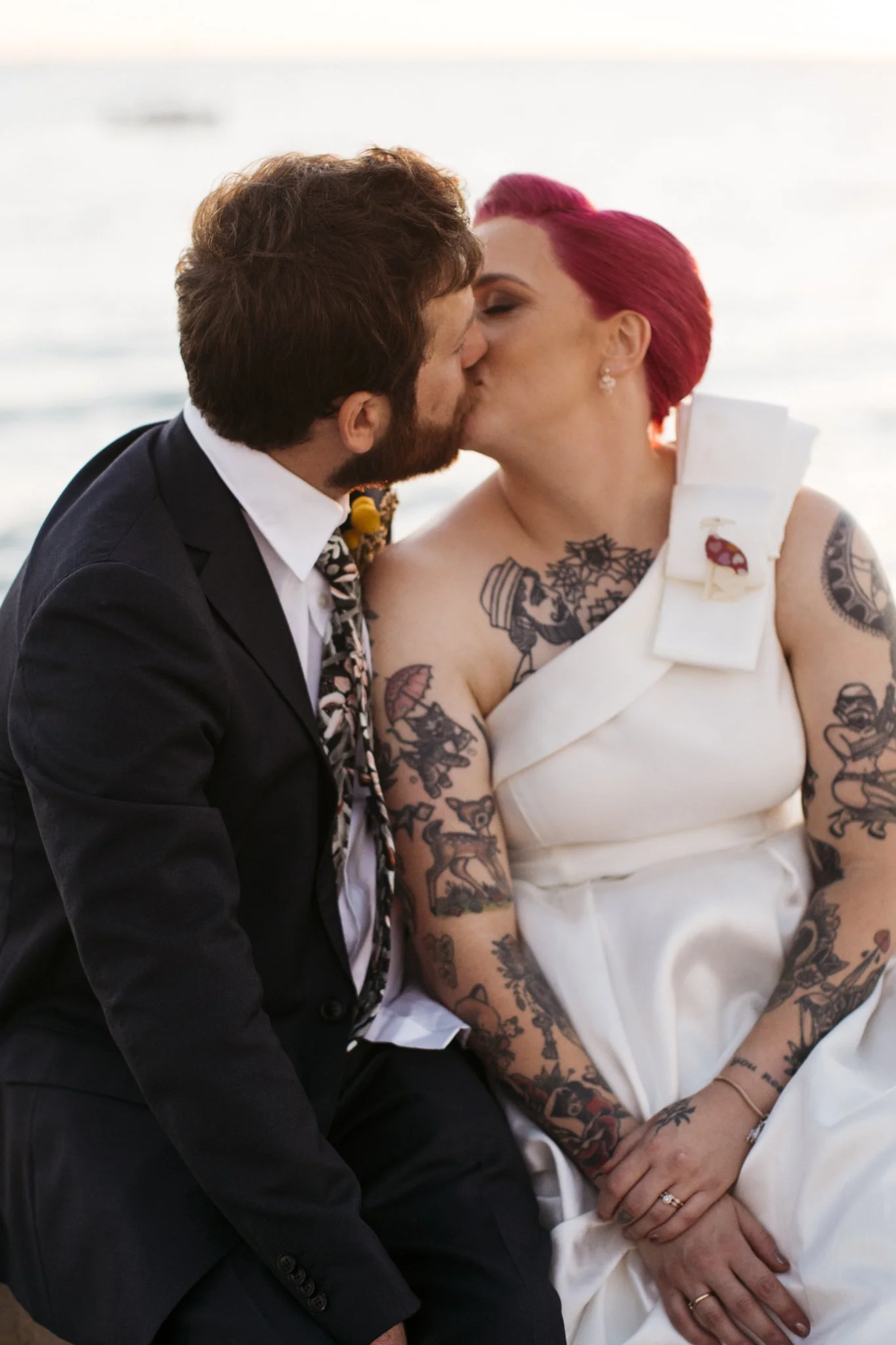 A couple in wedding attire sharing a kiss near a body of water, with the bride having bright pink hair and visible tattoos.