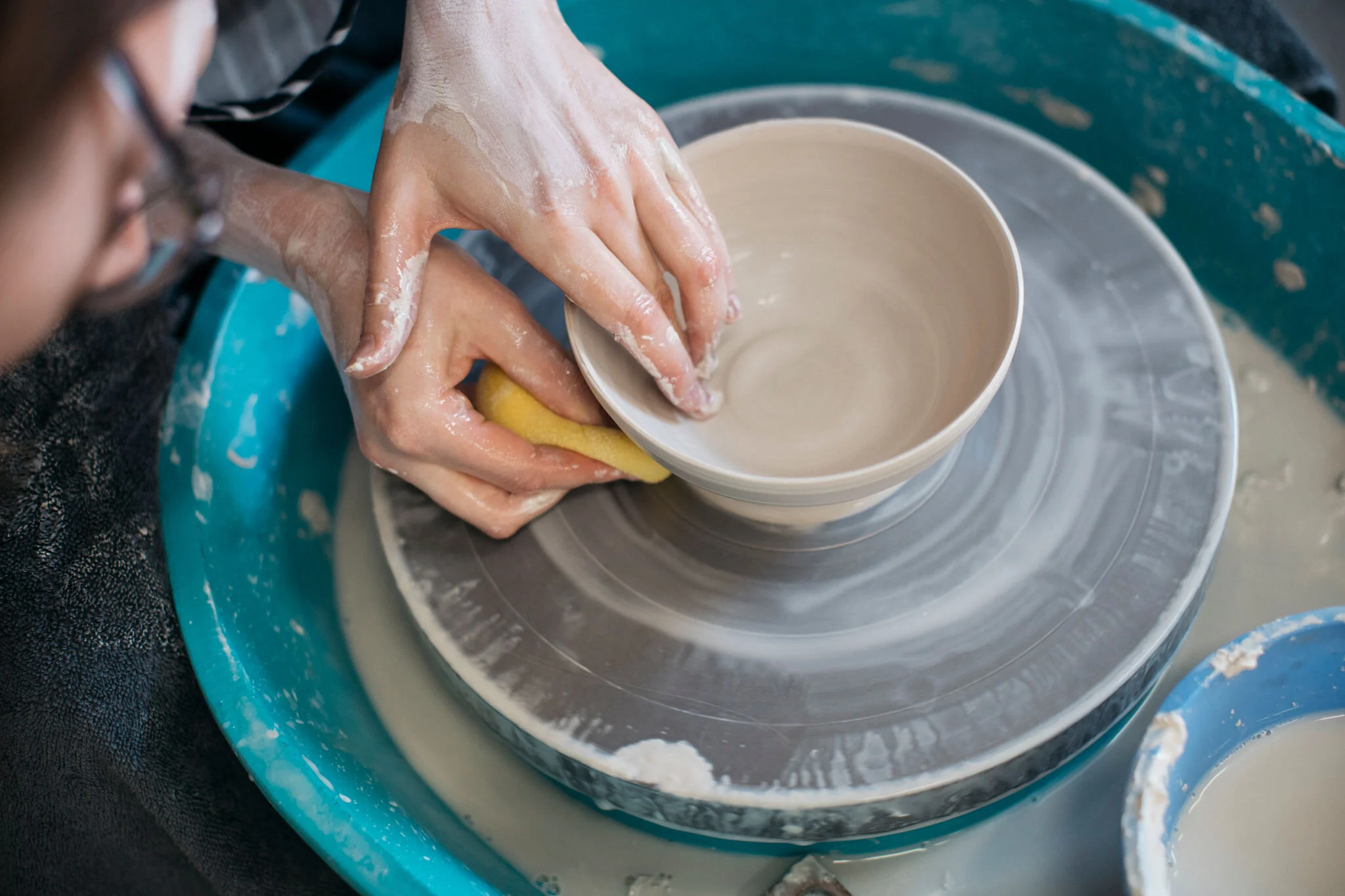 A person is using a sponge to smooth the inside of a ceramic bowl on a pottery wheel, with water and clay smeared on their hands.