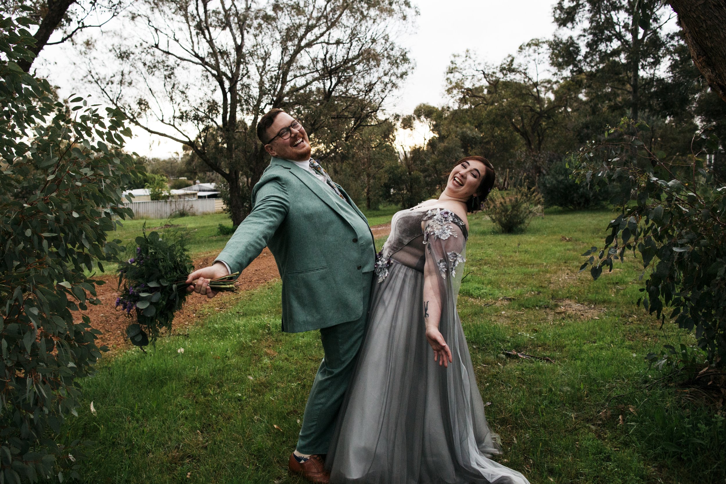 A couple dressed in formal attire laughing and posing playfully touching stomachs outdoors, with the man holding a bouquet of flowers on a grassy area surrounded by trees.