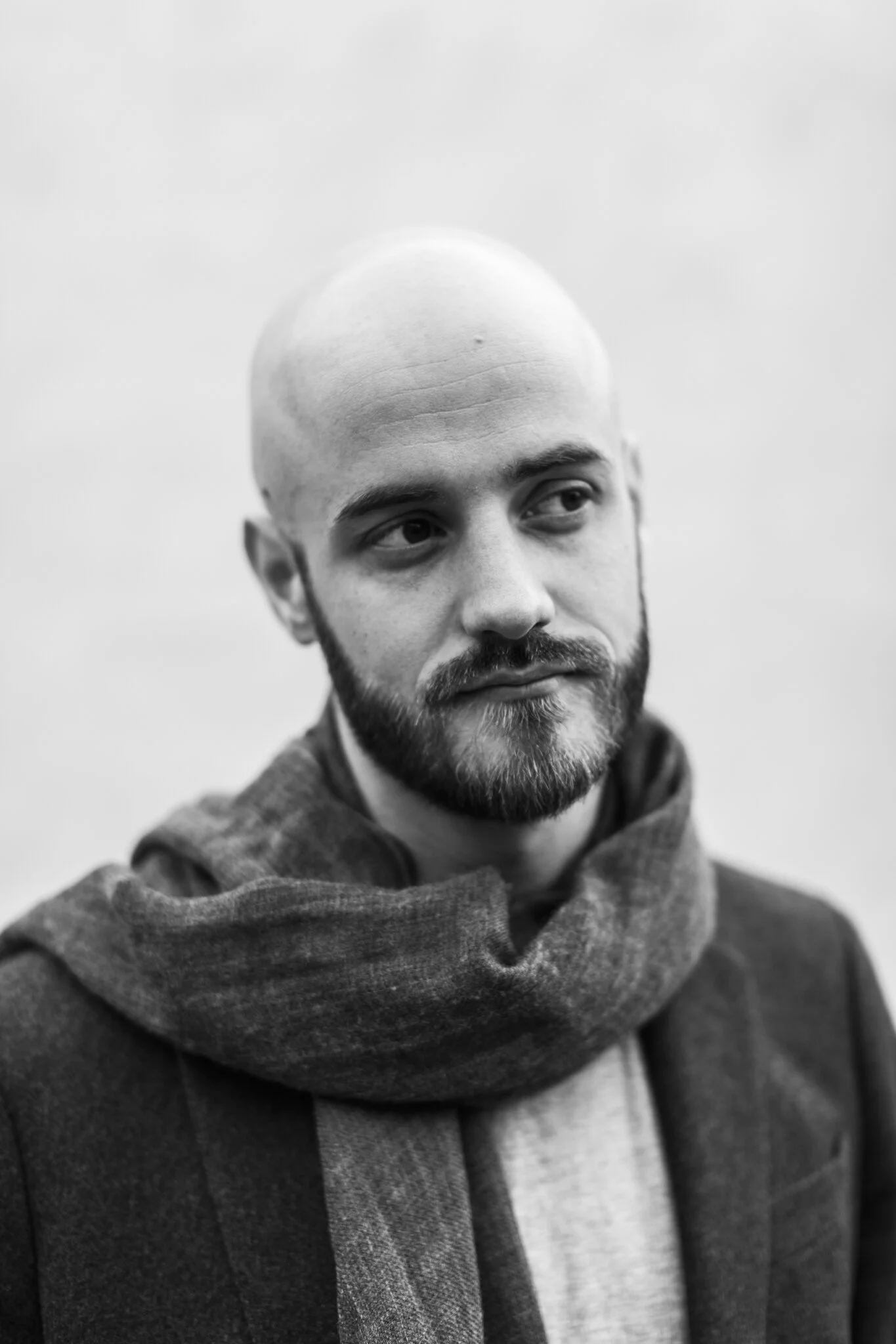 Black and white photo of a man with a shaved head, beard, and mustache, looking off to the side, wearing a scarf and coat.