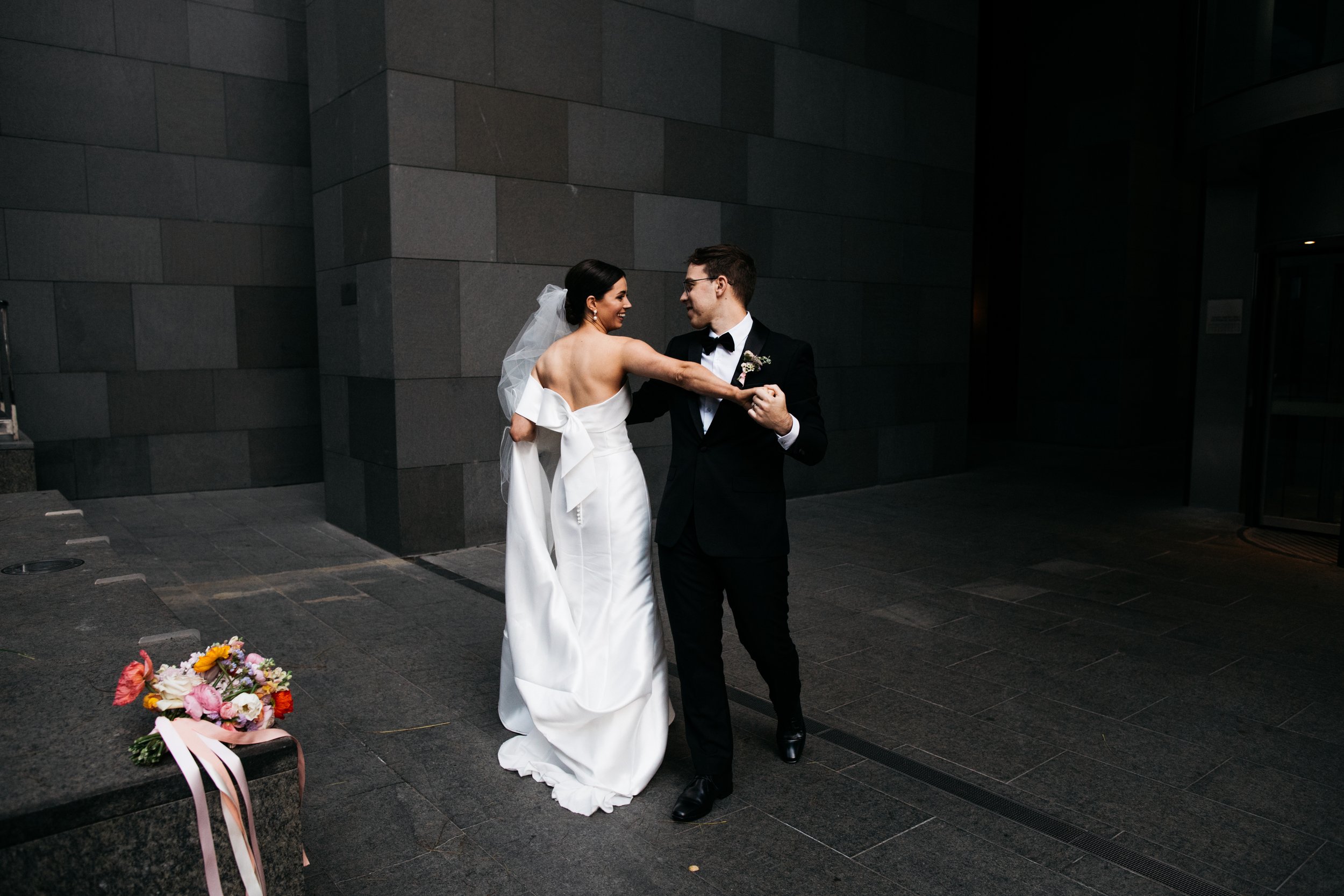 Bride and groom dancing together outside against a dark gray stone wall, bride in a white gown with off-the-shoulder sleeves, groom in a black suit with a bow tie, bouquet of flowers on the ground beside them.