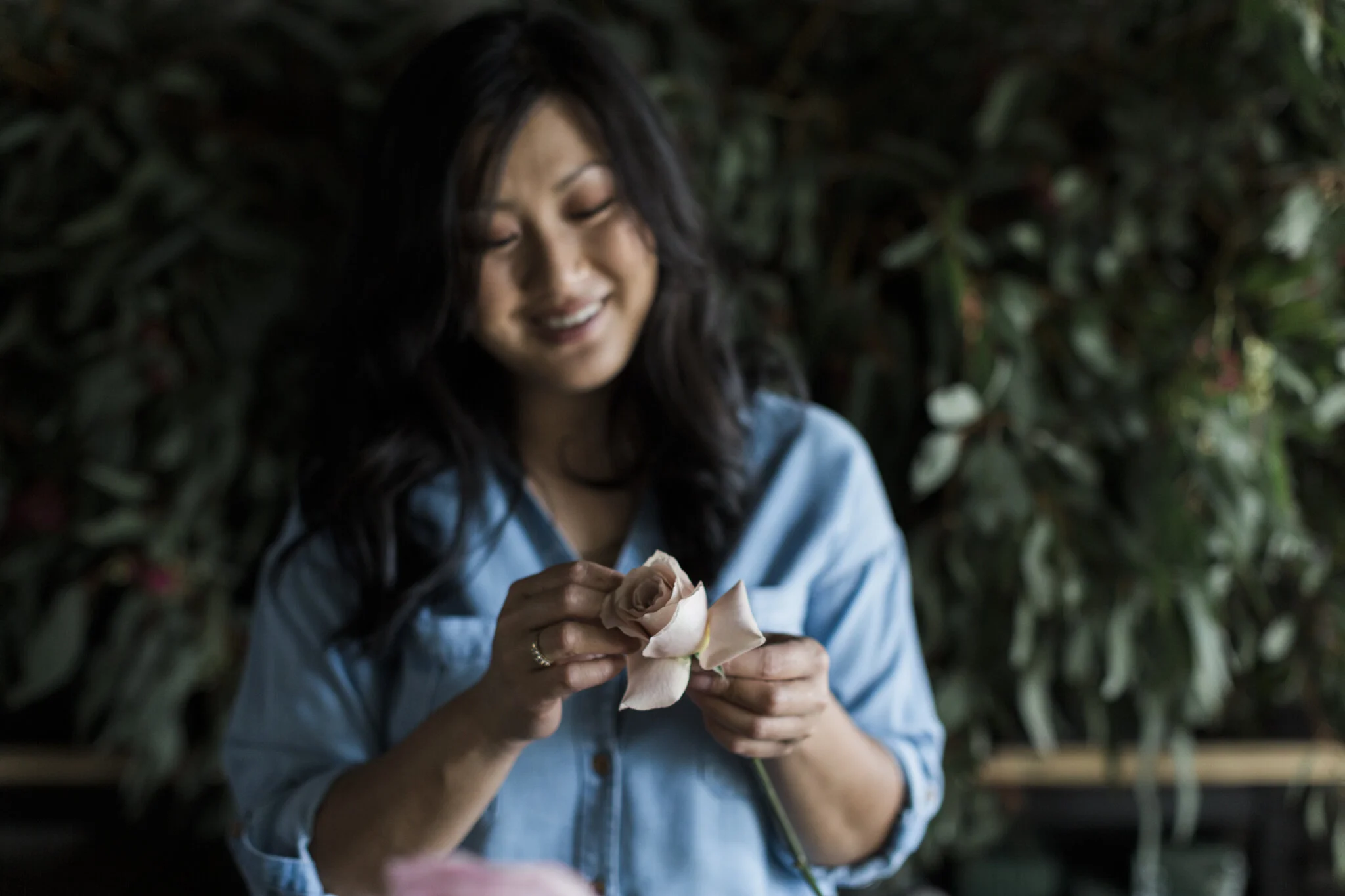 A woman smiling while holding a pink rose in her hands, with green foliage in the background.