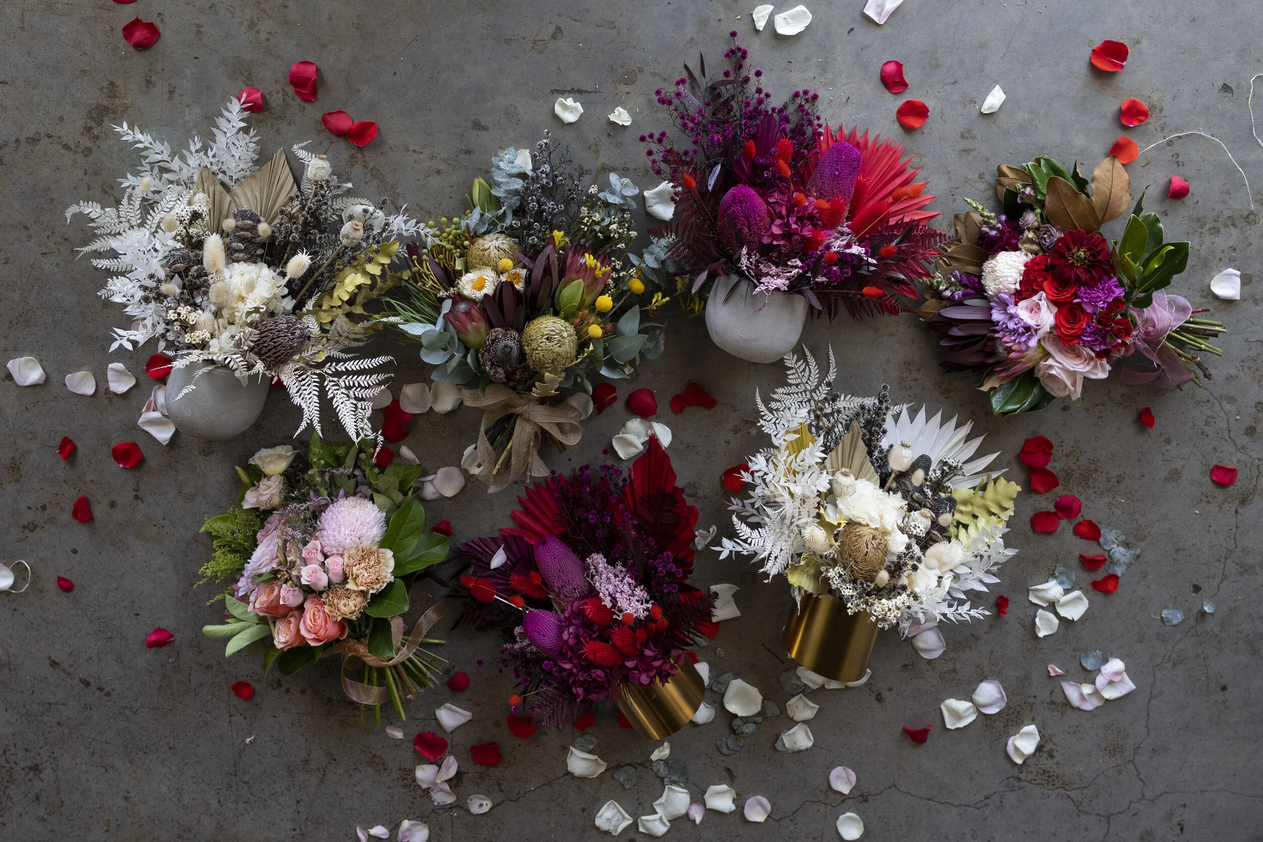 Five colorful flower bouquets arranged on a concrete floor, scattered with rose petals and small paper hearts.