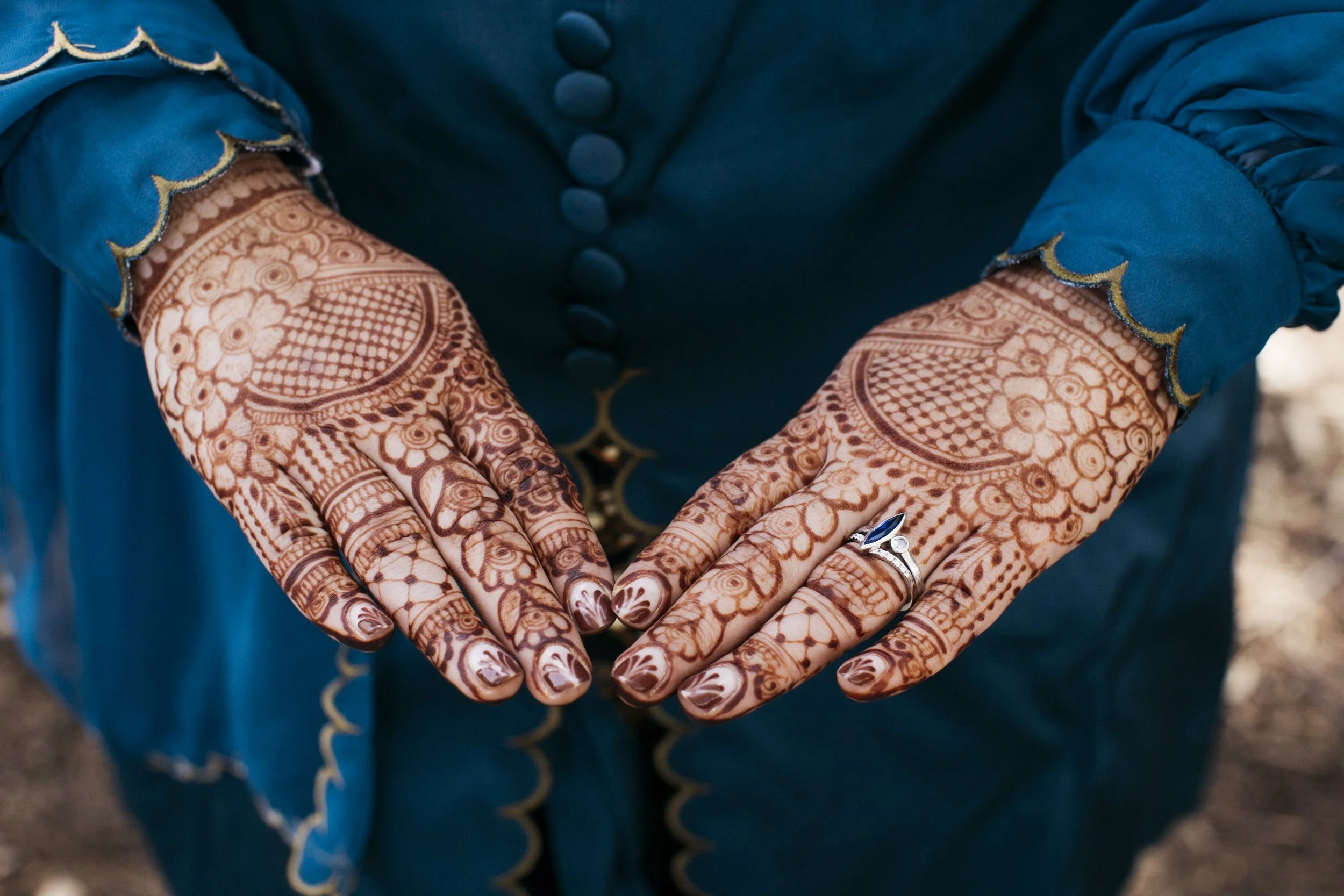 Close-up of hands with intricate henna designs, wearing rings, in blue traditional attire with gold accents.