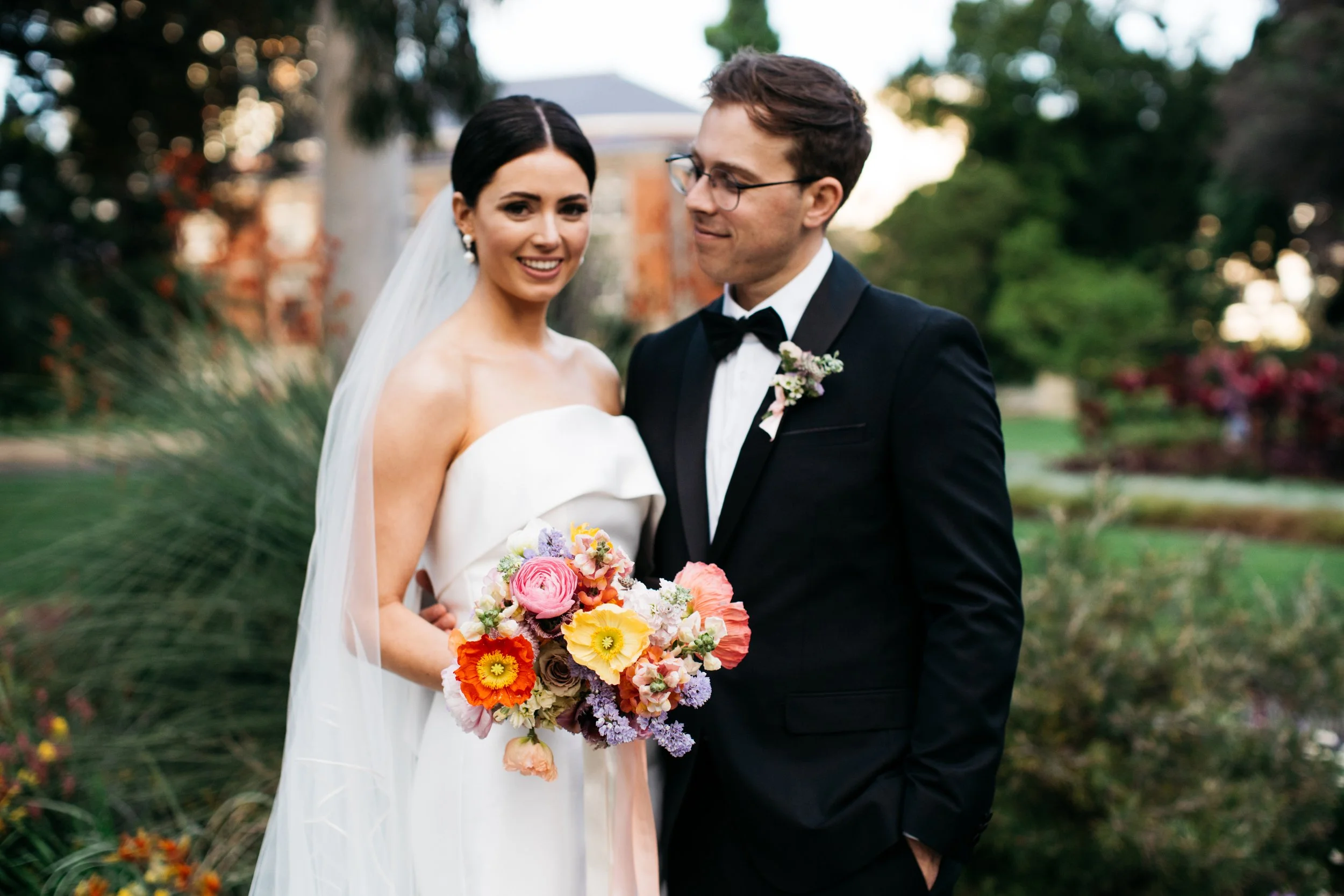 Bride and groom standing outdoors on their wedding day, smiling, with the bride holding a colorful bouquet of flowers, and the groom wearing a black tuxedo with a bow tie, in front of a lush green garden.