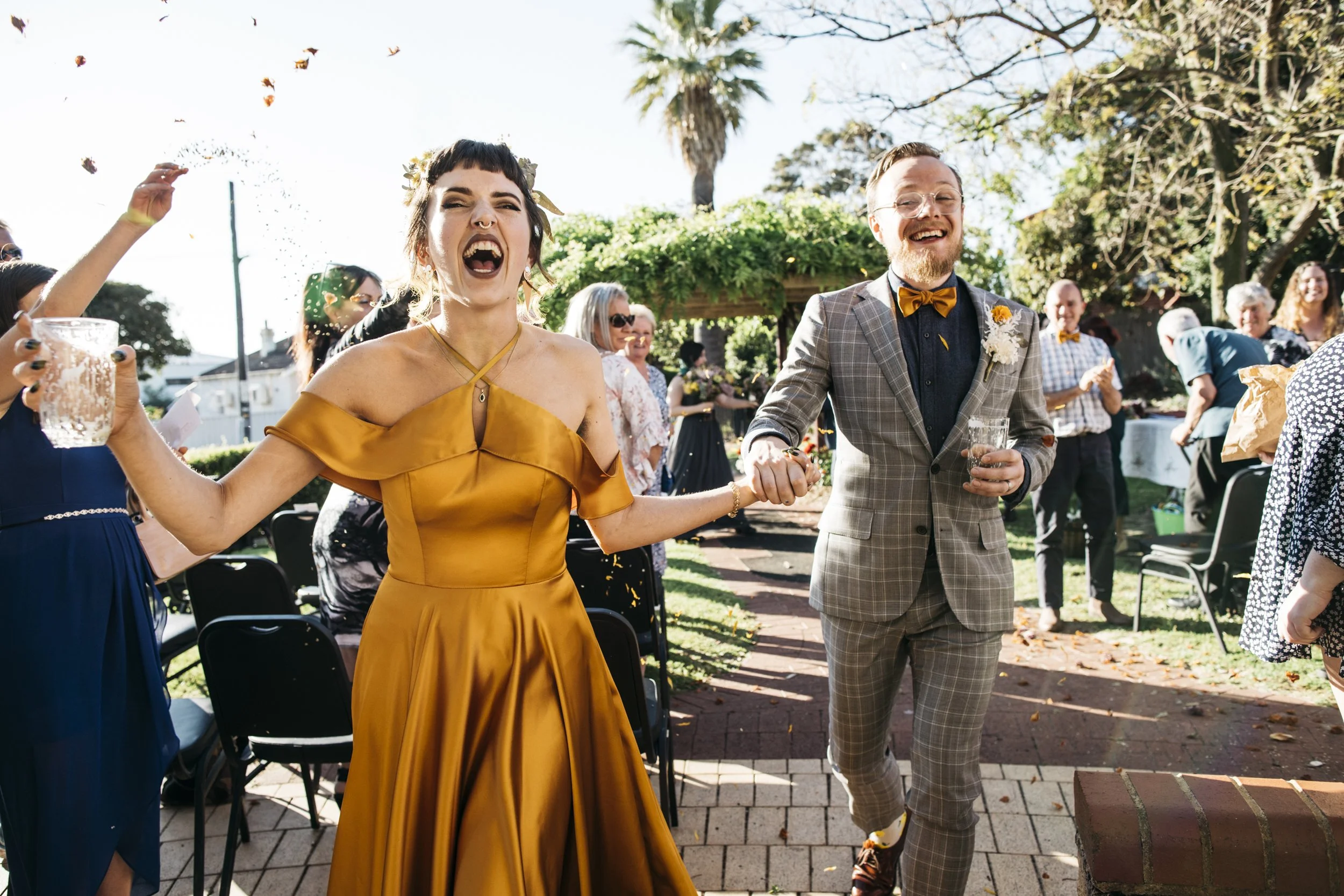 A bride and groom at their wedding celebration outdoors, holding hands and smiling. The bride is wearing a gold satin dress, and the groom is in a gray checkered suit with a bow tie. Guests are in the background, some cheering and holding drinks, with sunlight filtering through trees.