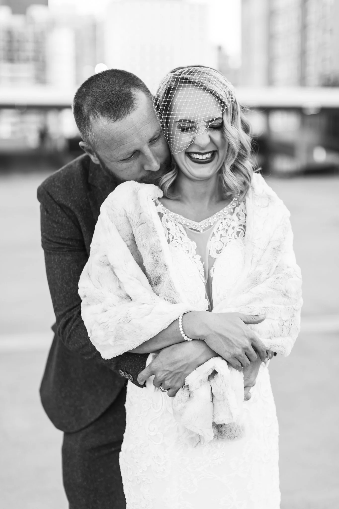 Black and white photo of a man and woman, with the man hugging the woman from behind, both smiling and laughing. The woman is wearing a lace dress, a fur wrap, and a small birdcage veil. The man is dressed in a suit. They appear to be celebrating a s