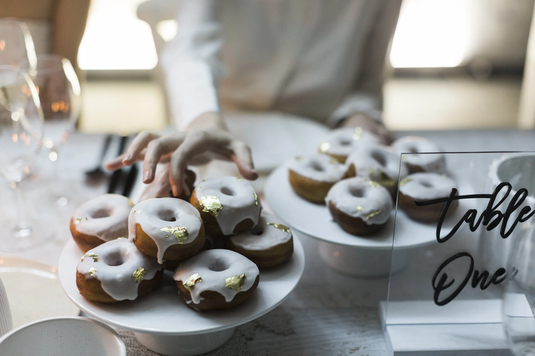 Plate of donuts with white icing and gold leaf decorations at a table, with a hand reaching for a donut.