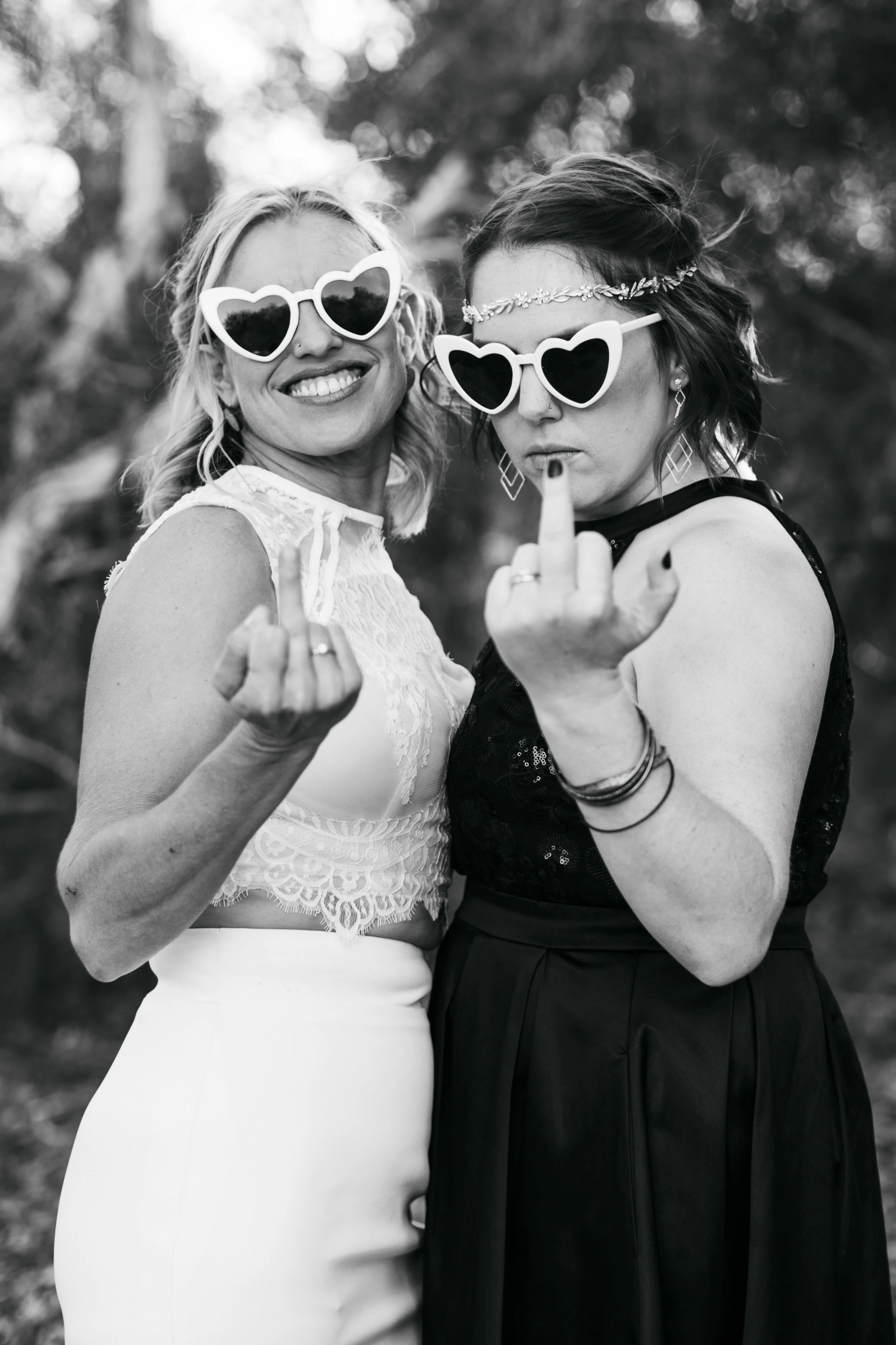 Two women wearing heart-shaped sunglasses showing middle fingers at the camera, one smiling and the other serious, outdoors with trees in the background.