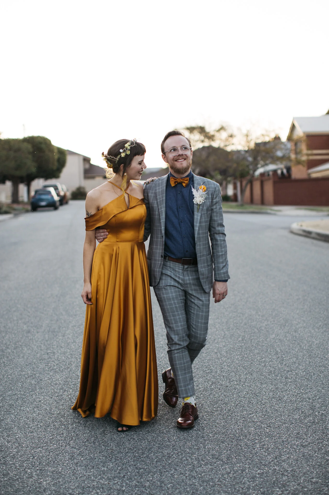 A couple walking arm in arm on a street during sunset, dressed in formal attire with the woman in a mustard yellow gown and the man in a plaid suit with a mustard bow tie and boutonniere.