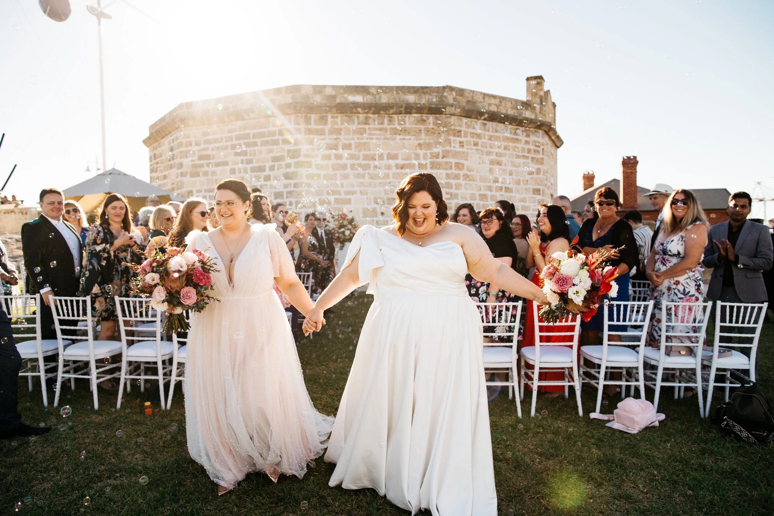 Two women in wedding dresses holding hands and walking on grass at an outdoor wedding reception, with guests seated on white chairs and celebrating in the background.