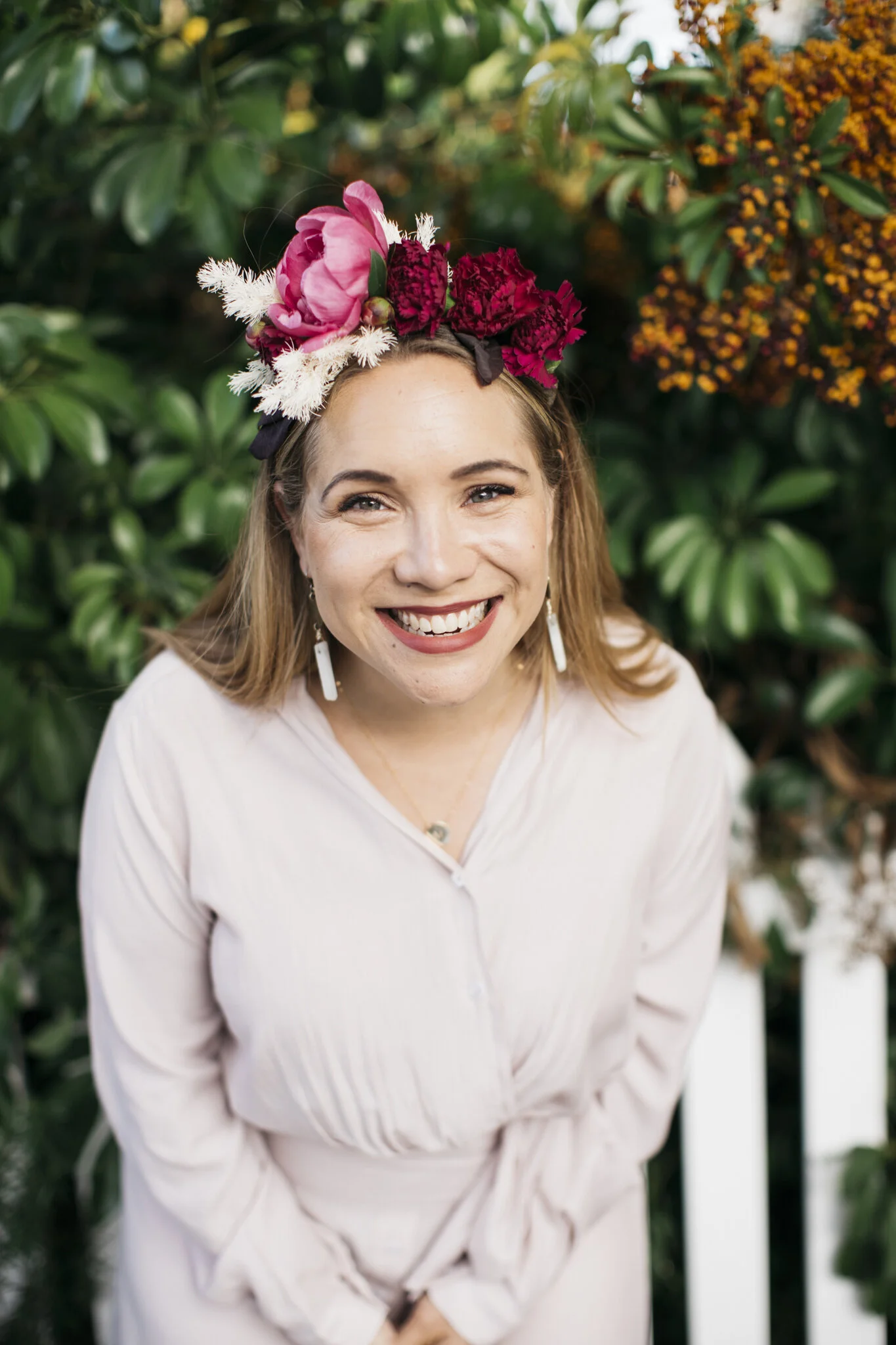 A smiling woman with light brown hair wearing a floral headband and white earrings, standing in front of green foliage and orange flowers.