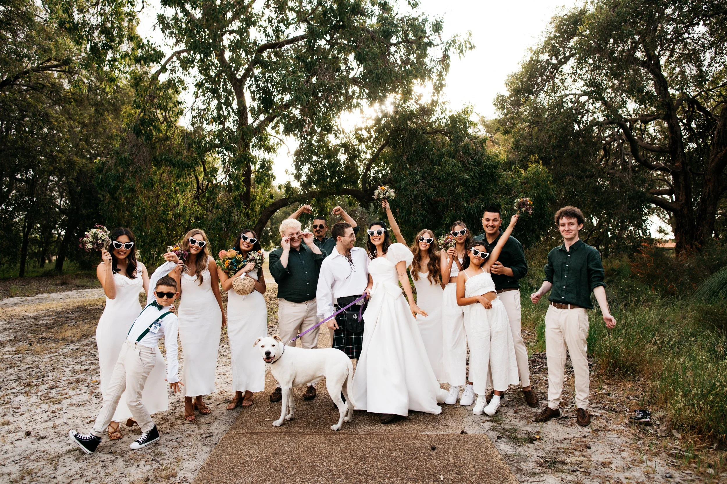 Group of people, including a bride and groom, celebrating outdoors with trees in the background. Many are wearing white clothing and heart-shaped sunglasses, holding bouquets, with a dog in front. The setting appears to be a wedding or celebration.