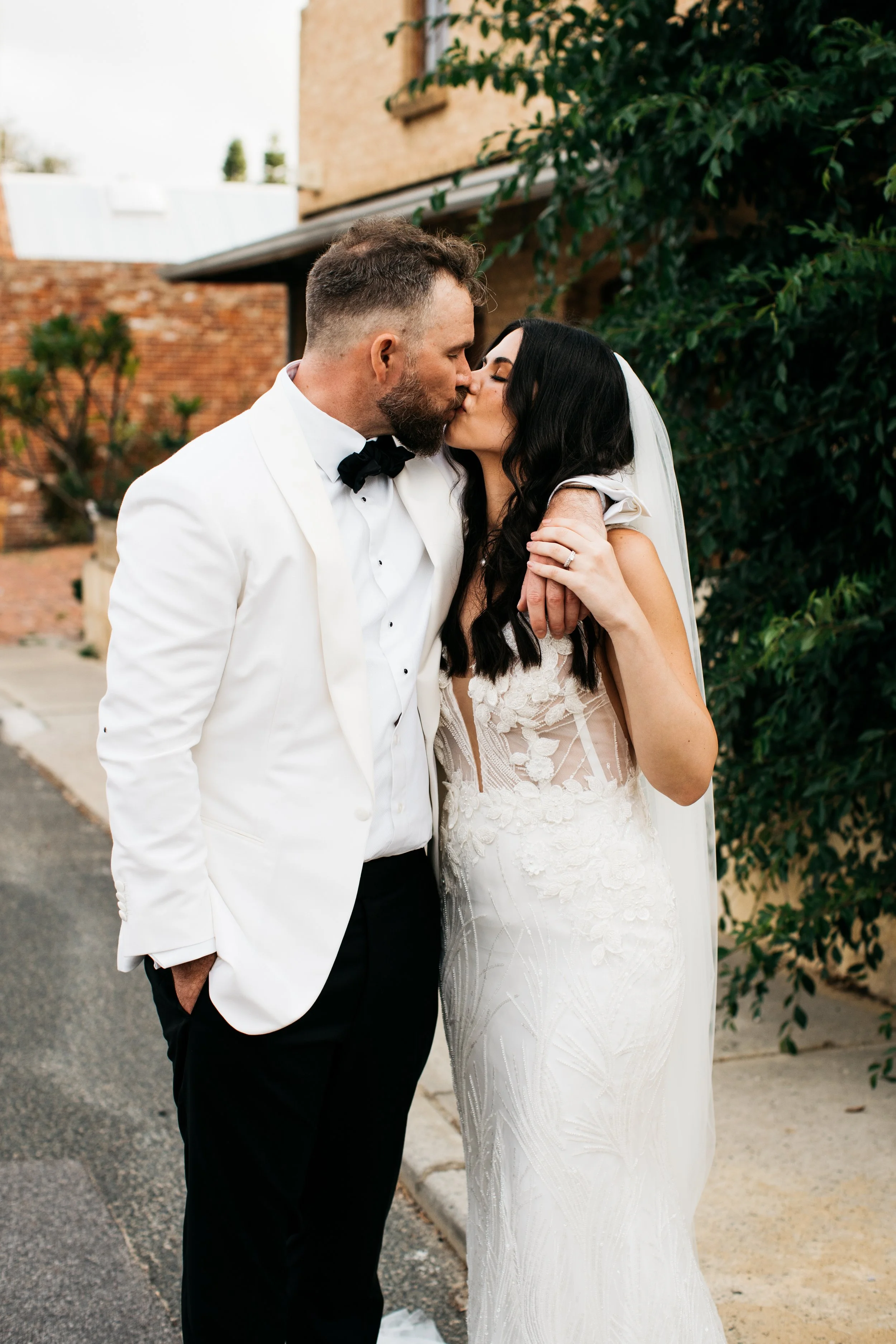 A bride and groom kissing outdoors on their wedding day, with the bride in a white lace wedding dress and veil, and the groom in a white tuxedo jacket and black pants, standing on a street near greenery and brick buildings.