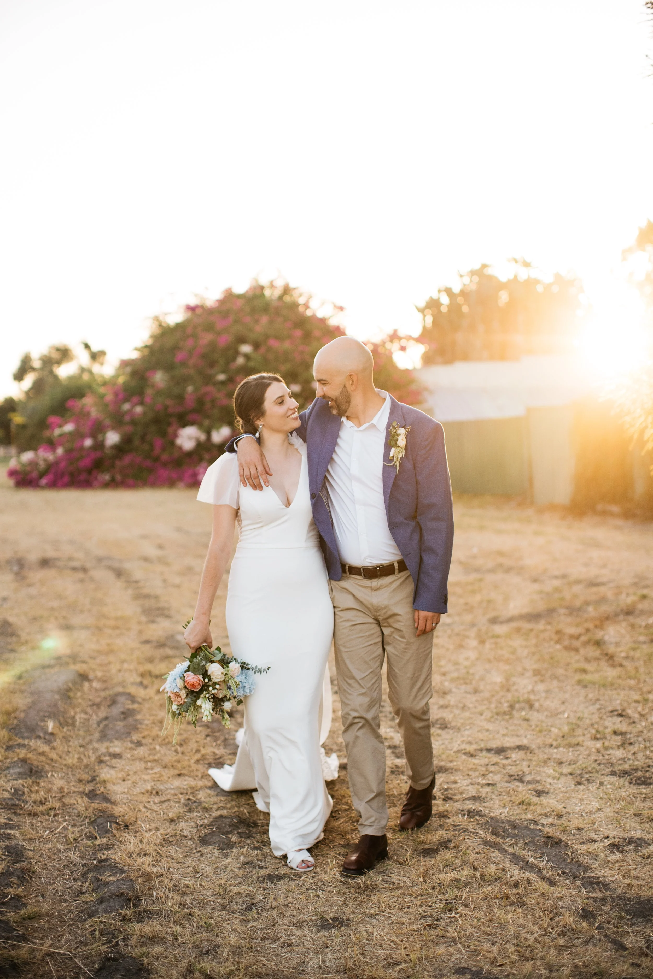 A newlywed couple walking outdoors during sunset, with the woman in a white wedding dress holding a bouquet and the man in a blue blazer and beige pants.