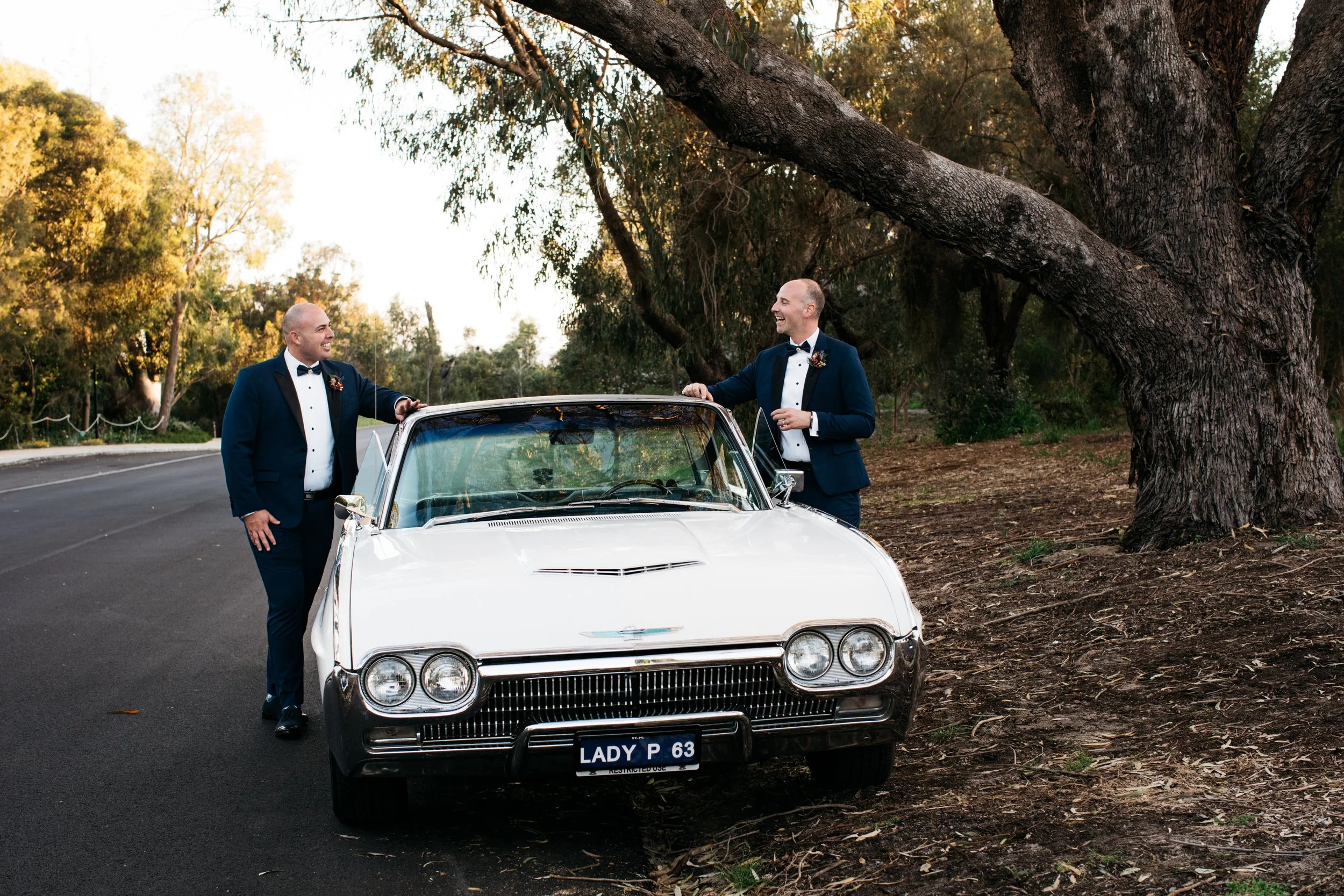 Two men in tuxedos with boutonnières, smiling and talking next to a vintage white car on a scenic road, surrounded by trees at sunset.
