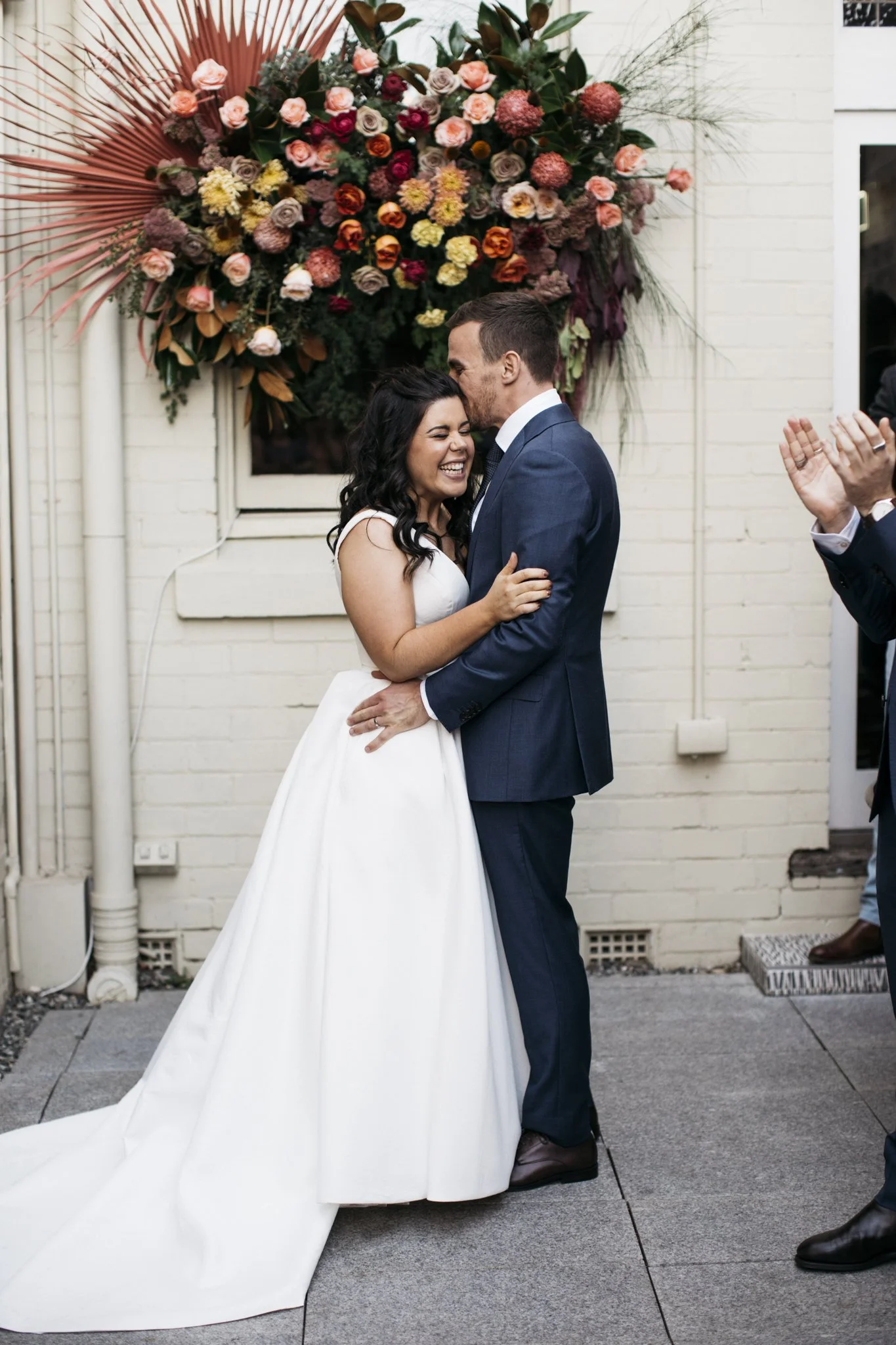 A bride and groom sharing a kiss and smiling during their wedding ceremony in front of a floral backdrop.