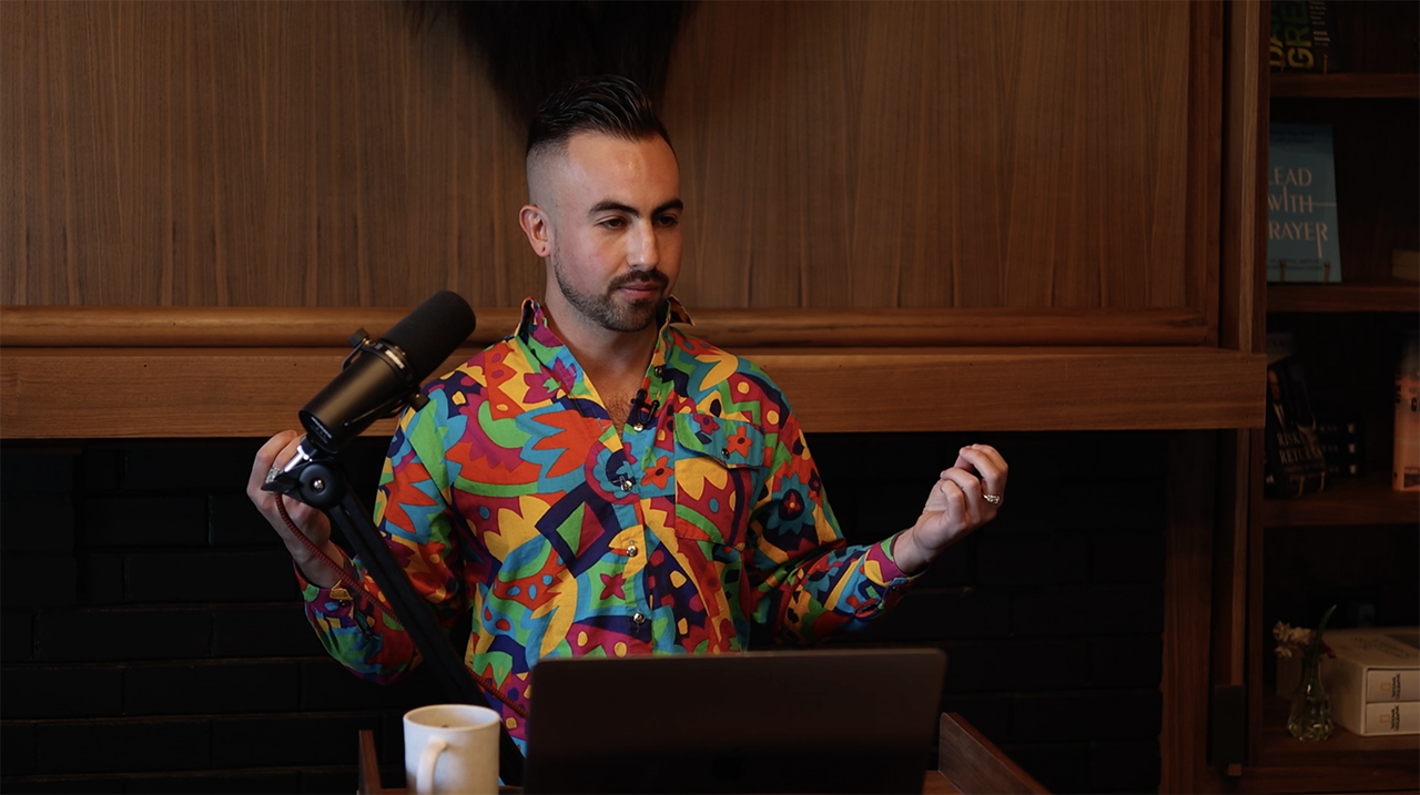 Dean Berlinerblau standing at a microphone during an oratory, with a laptop and mug, in a wood-paneled room lined with books.