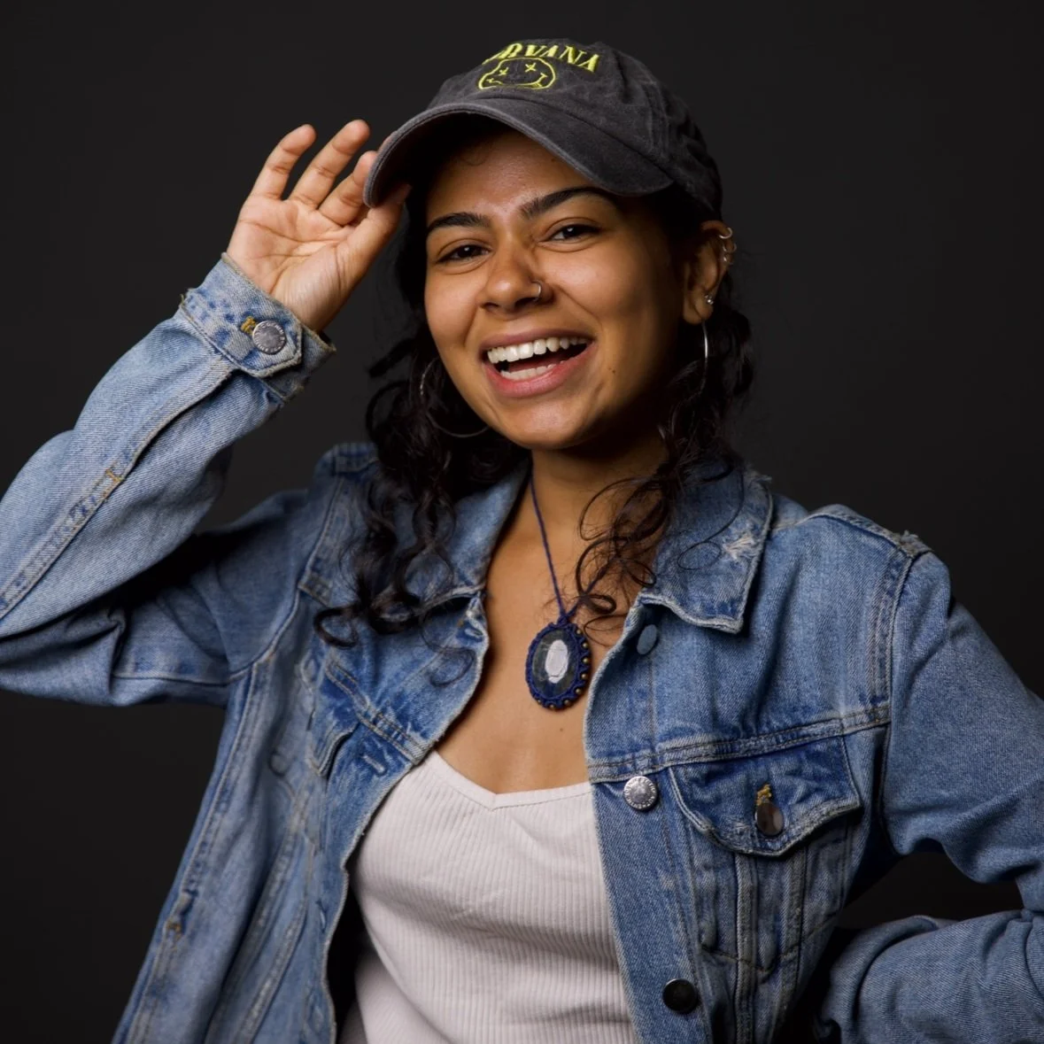 A smiling woman with curly hair, wearing earrings, a denim jacket, a white shirt, a blue pendant necklace, and a black cap with yellow accents, standing against a black background.