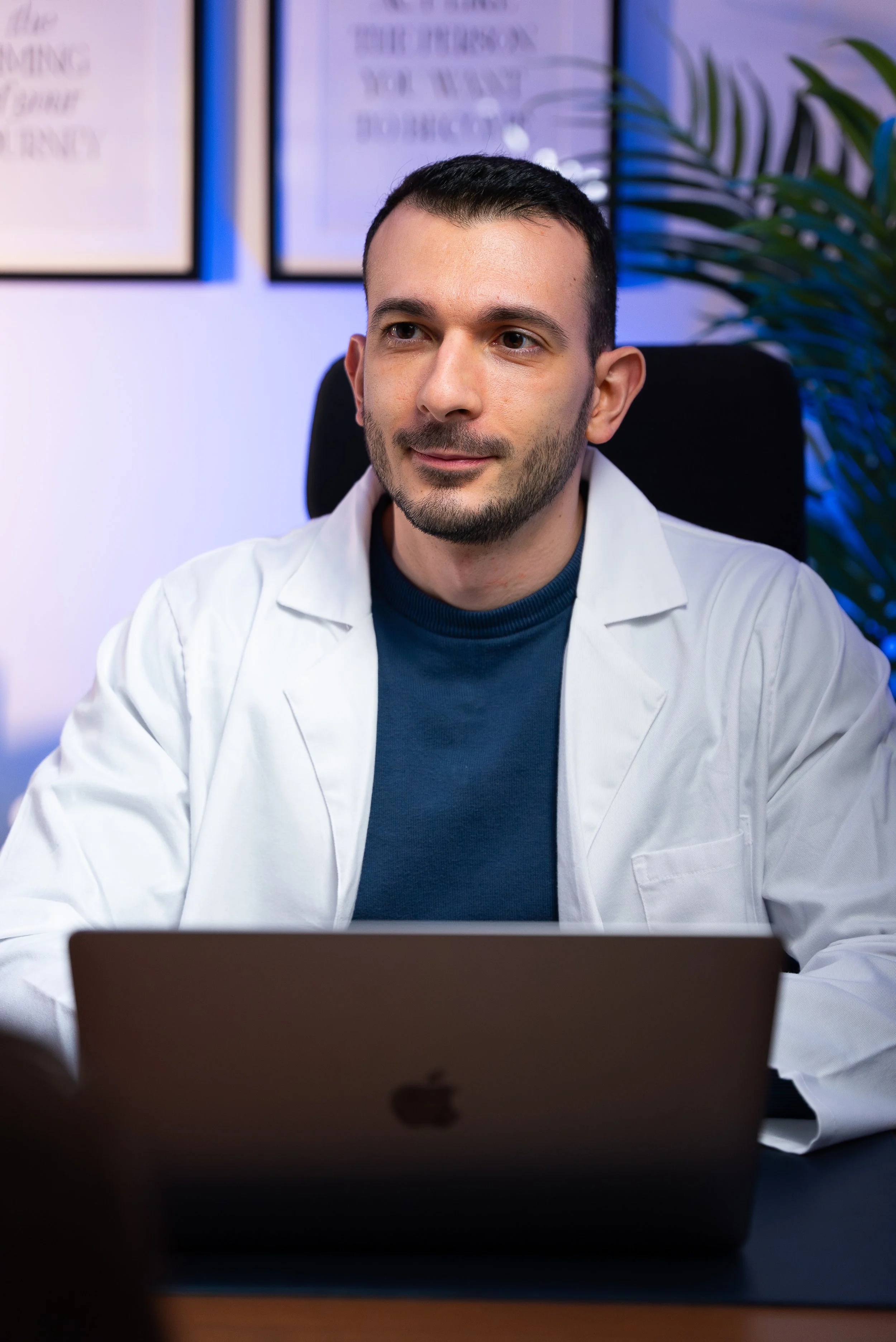 Un uomo con capelli corti e barba, indossa una giacca bianca e seduto davanti a un laptop, in un ufficio con pareti decorate e una pianta verde sullo sfondo.