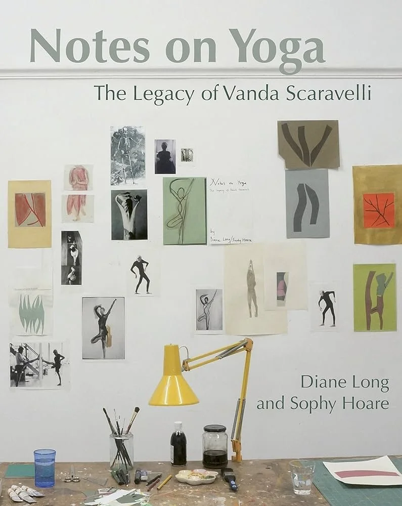 Wall display featuring the title 'Notes on Yoga: The Legacy of Vanda Scaravelli' with photos, sketches, and artwork of yoga poses, along with the names 'Diane Long and Sophy Hoare.' A yellow desk lamp and art supplies are on the table in front.