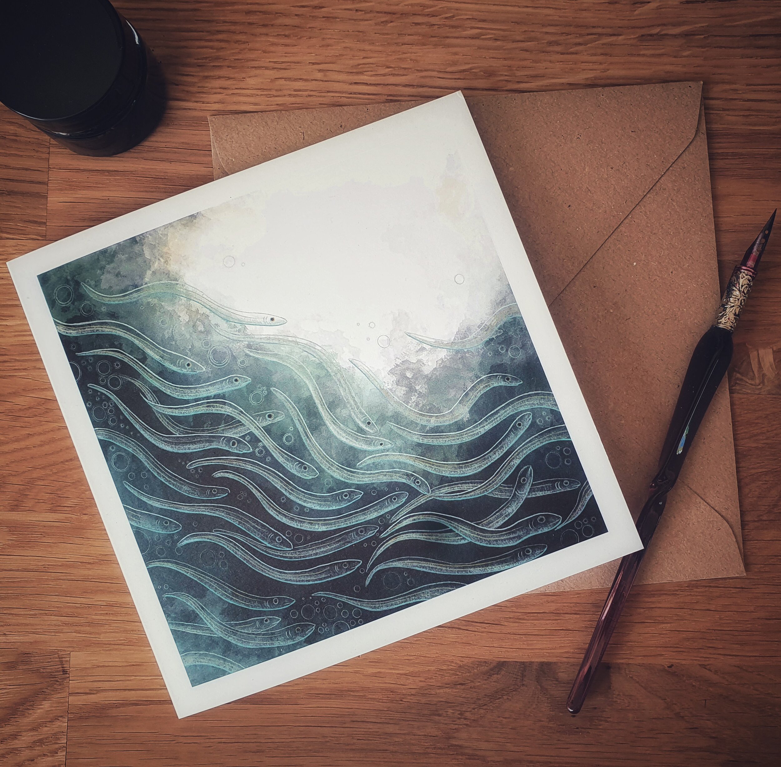 A greetings card with a brown envelope with a pen laying on a desk. Design on card shows a large group of glass eels swimming through deep turquoise water.