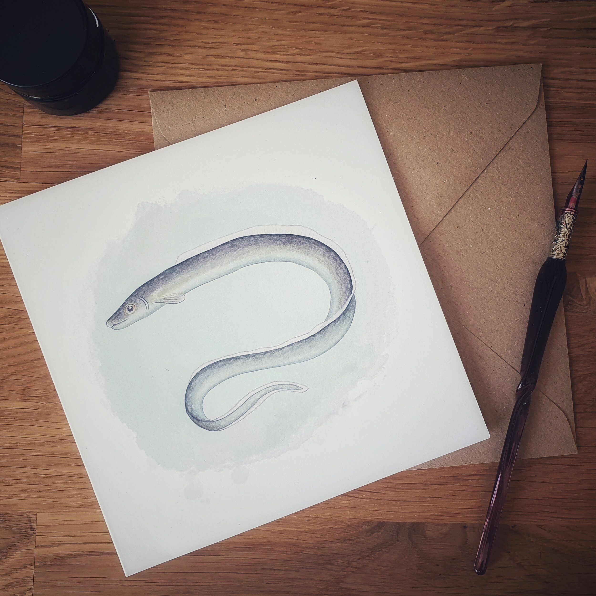 A greetings card with a brown envelope with a pen laying on a desk. Design on card shows Silver eel painted digitally in shades of blue and grey.