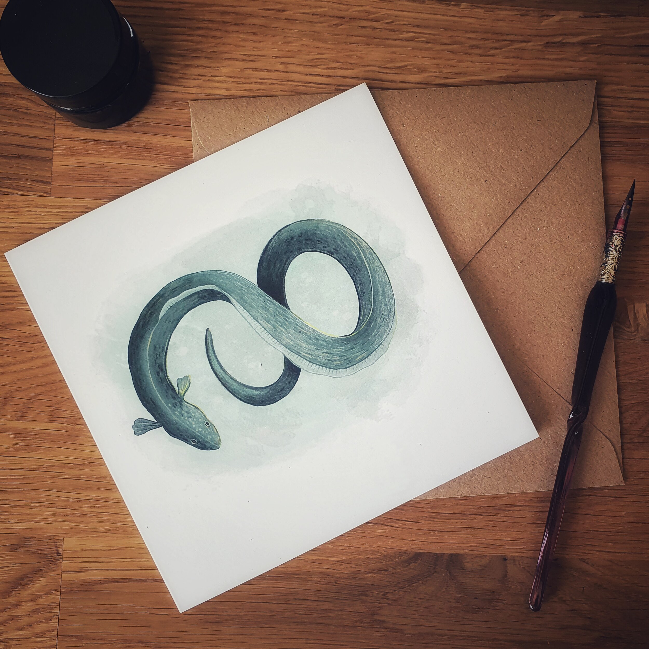 A greetings card with a brown envelope with a pen laying on a desk. Design on card shows a green eel in the shape of a figure of eight or infinity sign on a watercolour style background.