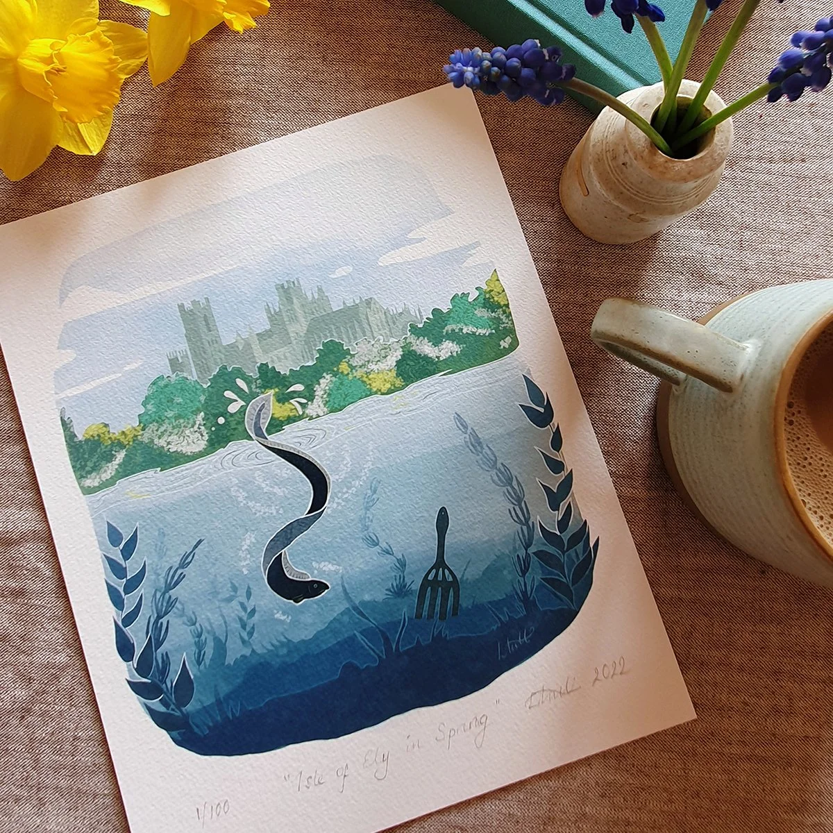 Giclee print of Ely Cathedral on a spring day with the river in the foreground where an eel swims next to an eel gleave. Print lays on linen cloth with flowers, books and coffee.