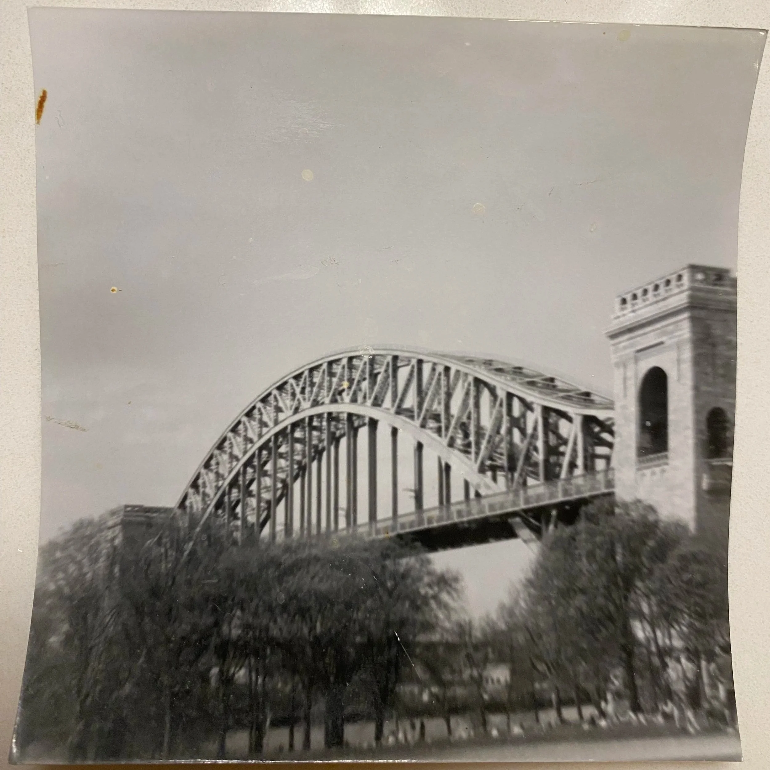 Black and white photo of the Hell Gate bridge with a tower on each side, spanning over trees in a park setting.