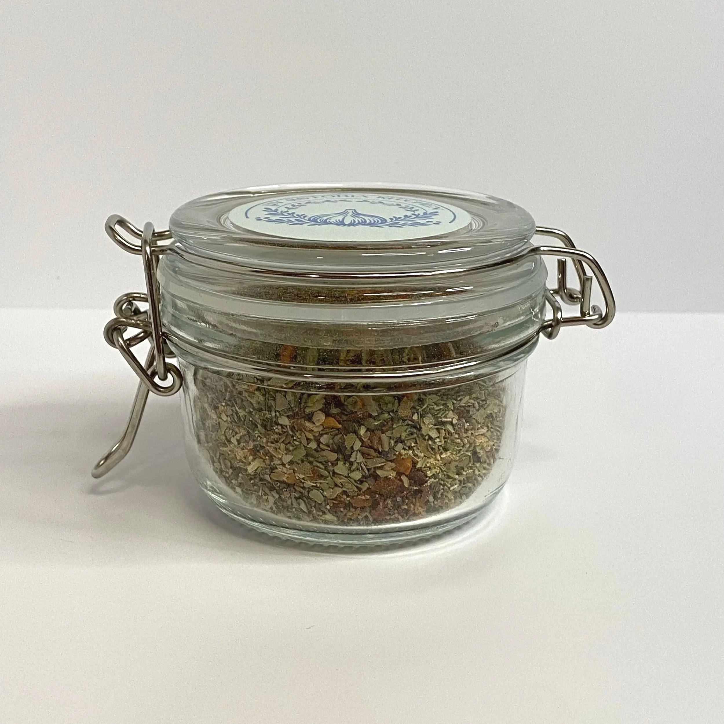Small glass jar with a metal clasp, filled with a dried herb mixture, sitting on a white surface against a white backdrop.