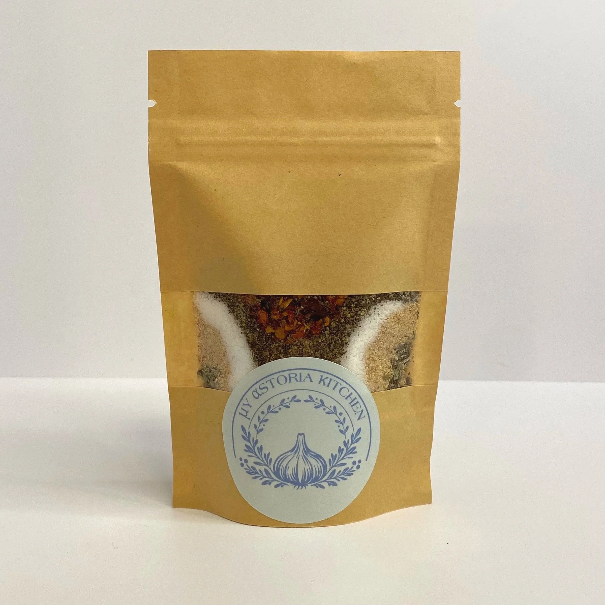 Brown paper bag with a clear window showing a mixture of spices and dried herbs, with a white circular label featuring a garlic illustration and the text "MY ASTORIA KITCHEN."