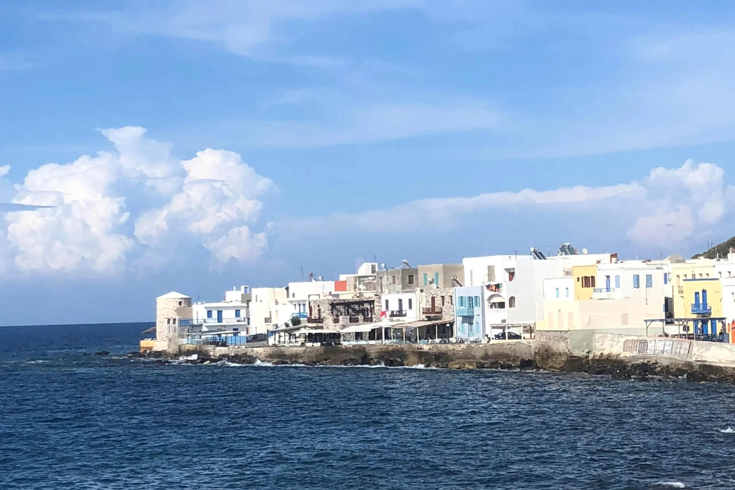 A coastal scene with a row of white and pastel-colored buildings along the shoreline under a partly cloudy sky, with the ocean in the foreground.
