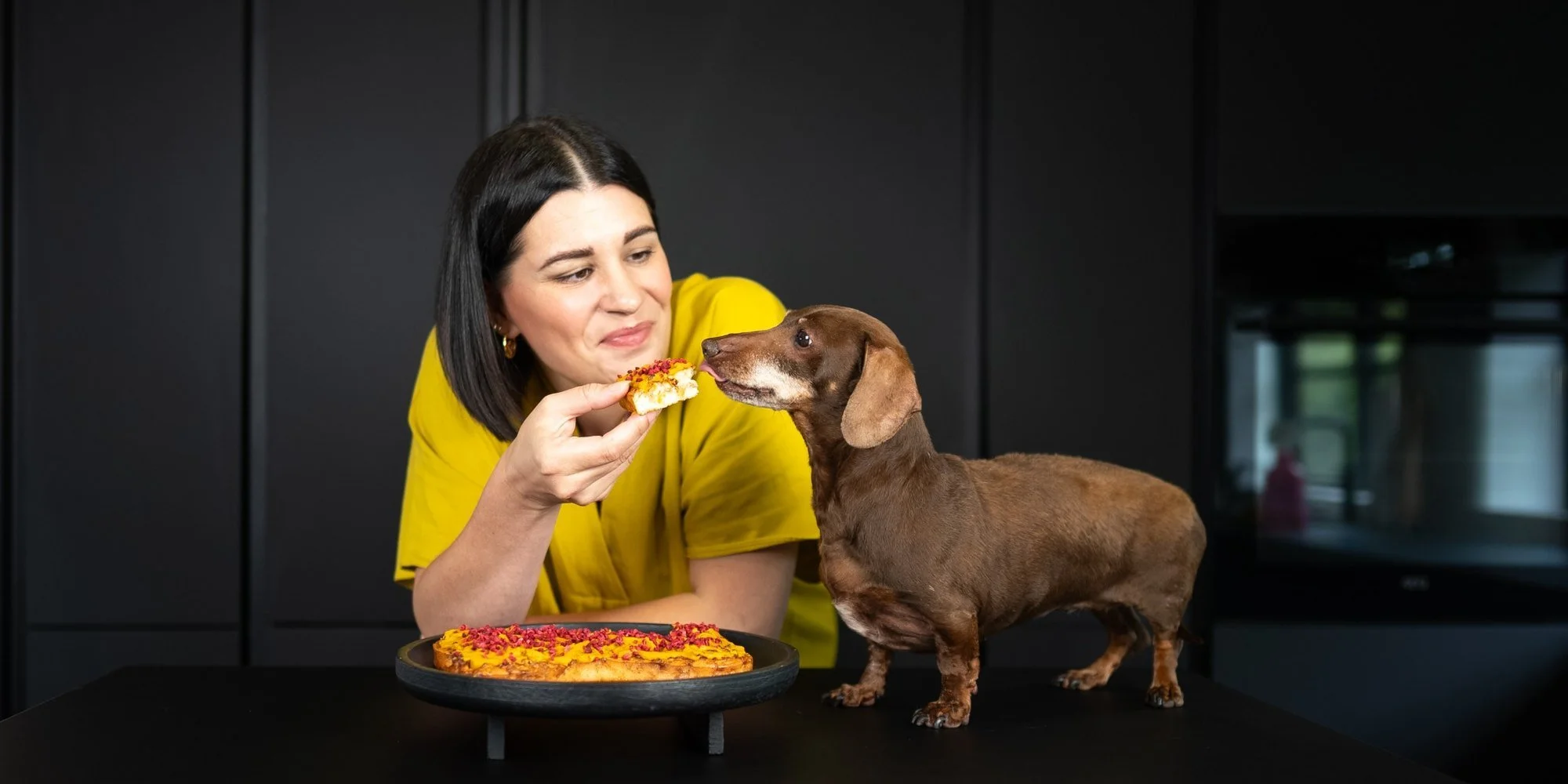 Woman sharing a homemade dog birthday cake with a dachshund in a kitchen