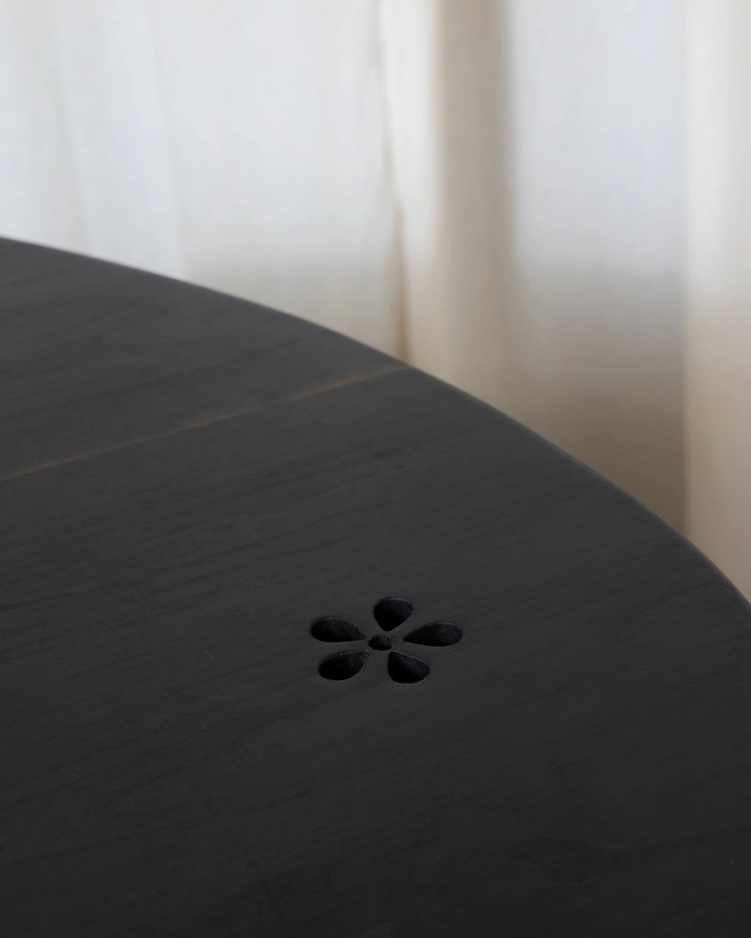 Close-up of a dark wooden surface with a carved flower pattern near the bottom center, background shows a white wall with soft shadows and sunlight.