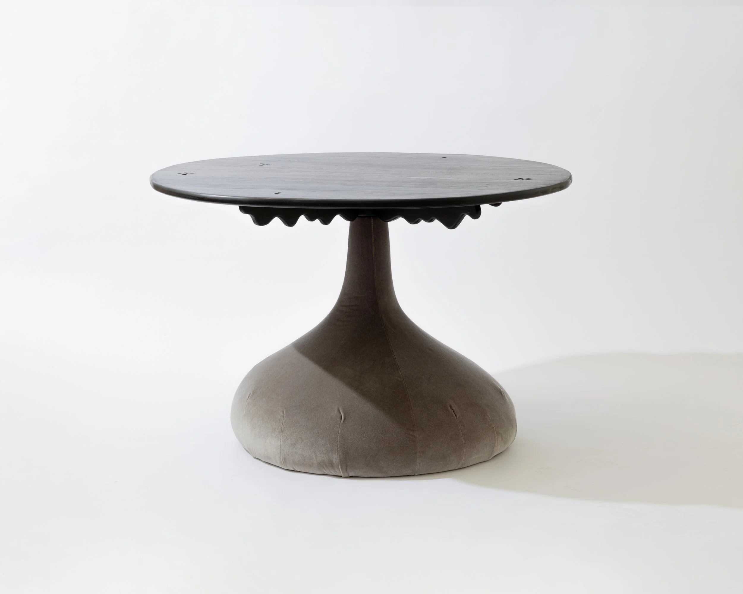 A round wooden top table with a single pedestal base, set on a pillow-shaped cushion