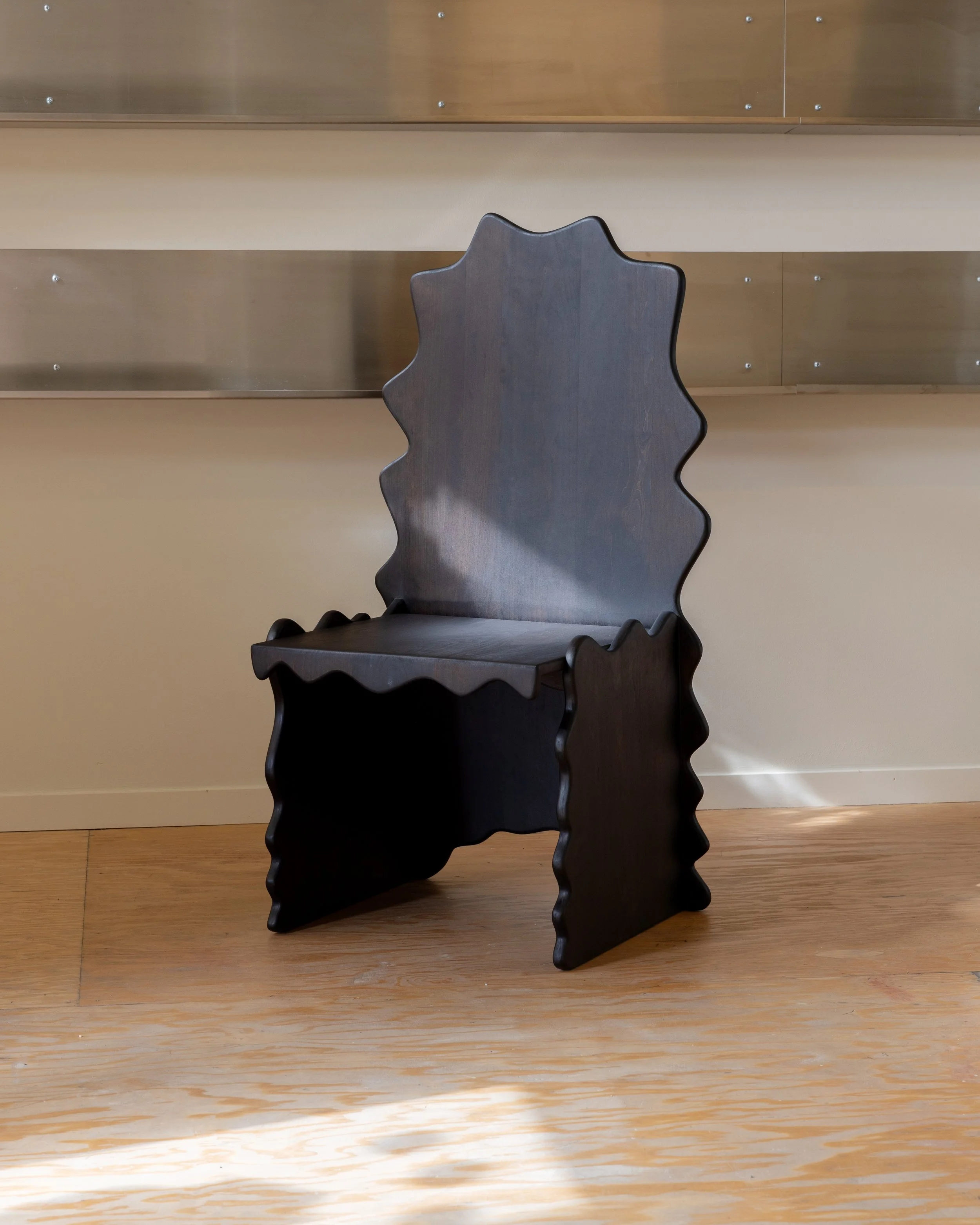 A dark wooden chair with a scalloped top and a carved wavy design on the backrest, placed on a light wooden floor against a beige wall with horizontal wooden paneling.