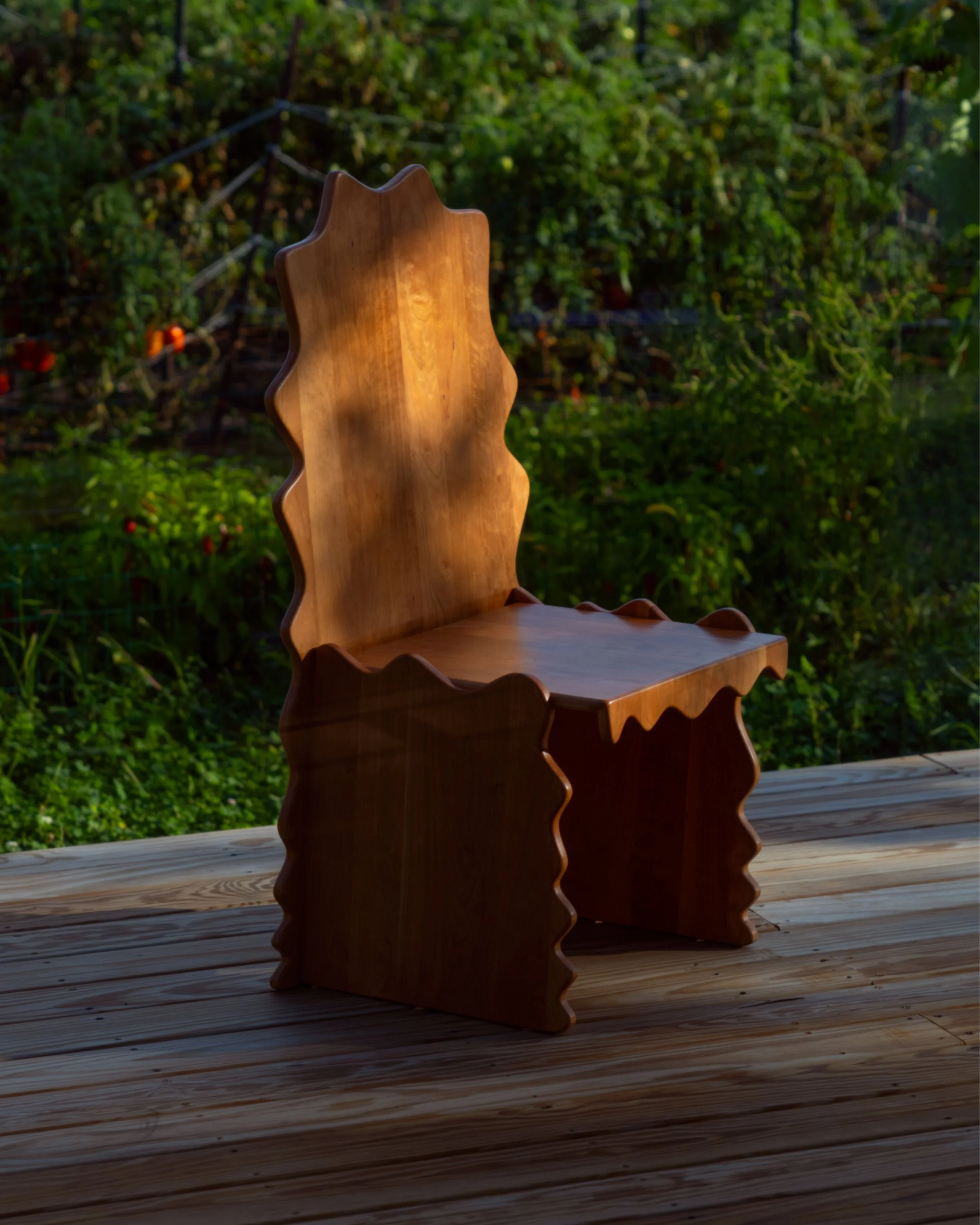 A wooden outdoor chair with a backrest and seat, positioned on a wooden deck with green foliage and garden in the background, illuminated by sunlight.