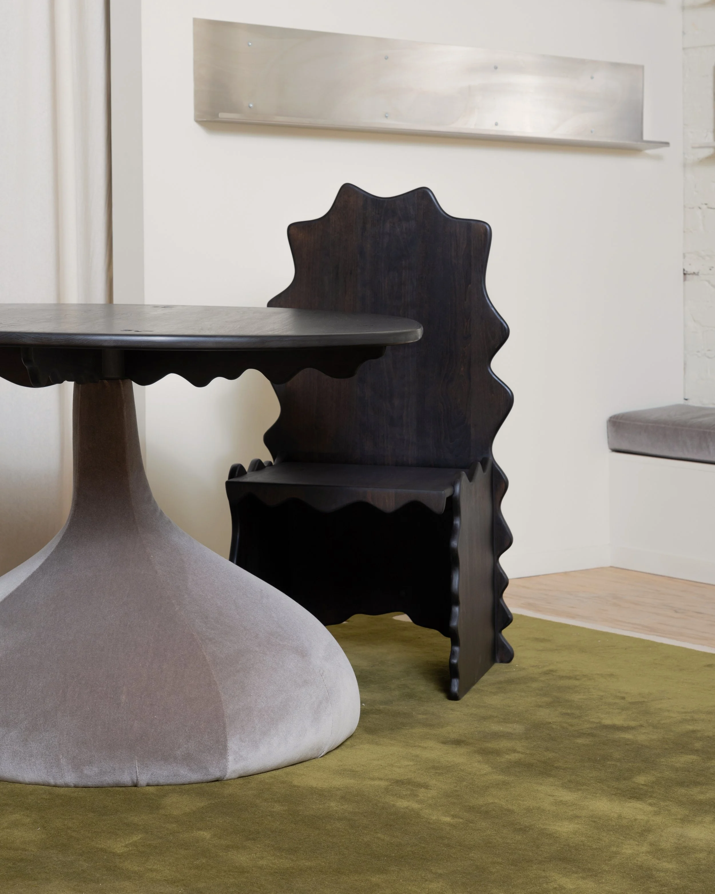 A close-up of a modern, abstract interior space with a dark wooden chair featuring a scalloped edge, a smooth gray ottoman, and a round green carpet on a wooden floor. Background includes a white wall with a light-colored abstract shelf.